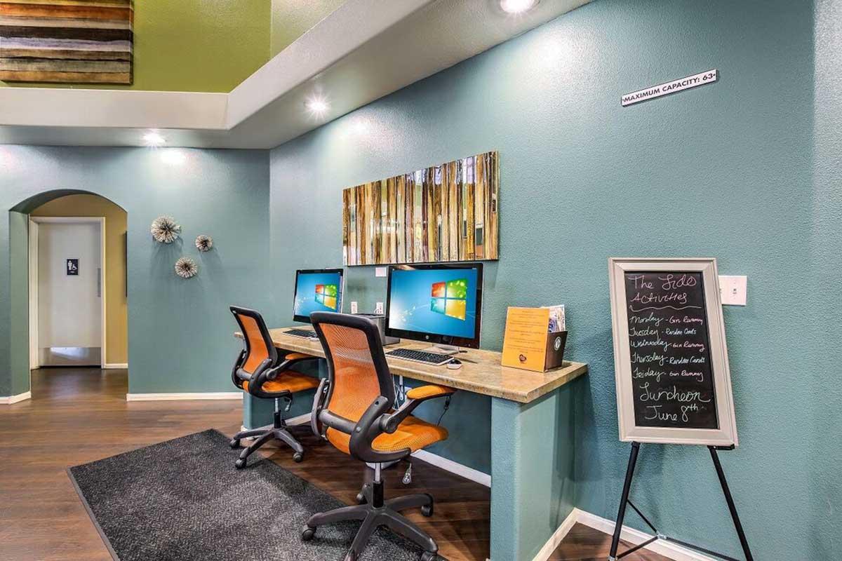 A modern office space featuring two computers on a wooden desk with orange chairs. The walls are painted in soft teal, and there are decorative wall accents, including a large mirror. A chalkboard lists upcoming events, and a sign indicates a maximum capacity. The overall atmosphere is bright and inviting.