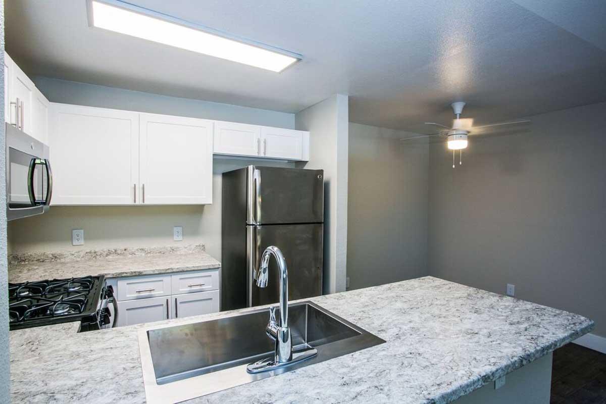 Modern kitchen featuring white cabinets, a stainless steel refrigerator, and a gas stove. The countertops are made of granite, with a large sink on the island. A ceiling fan is mounted in the corner, and the space is well-lit with overhead lighting. The walls are painted in a neutral color.