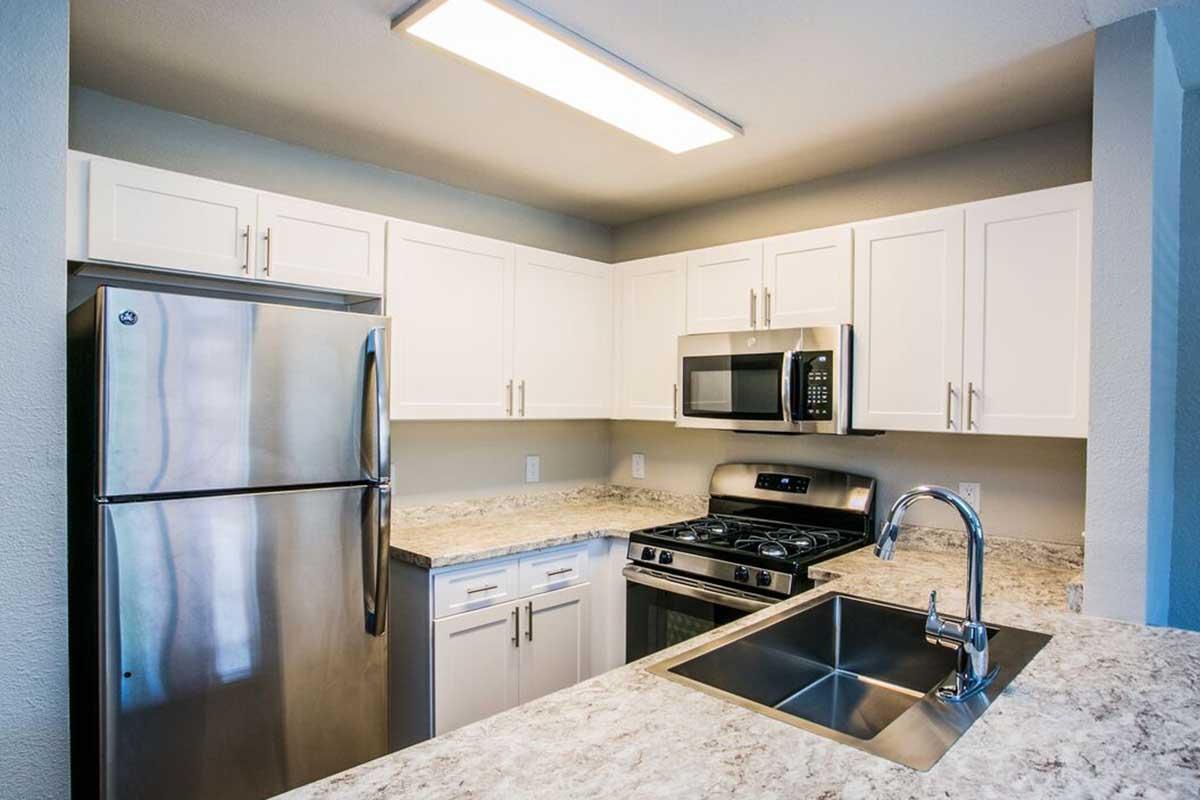 Modern kitchen featuring white cabinets, stainless steel appliances including a refrigerator, microwave, and gas stove. A granite countertop is visible with a built-in sink. Bright overhead lighting creates an inviting atmosphere. The walls are painted in a neutral tone, enhancing the overall aesthetic.