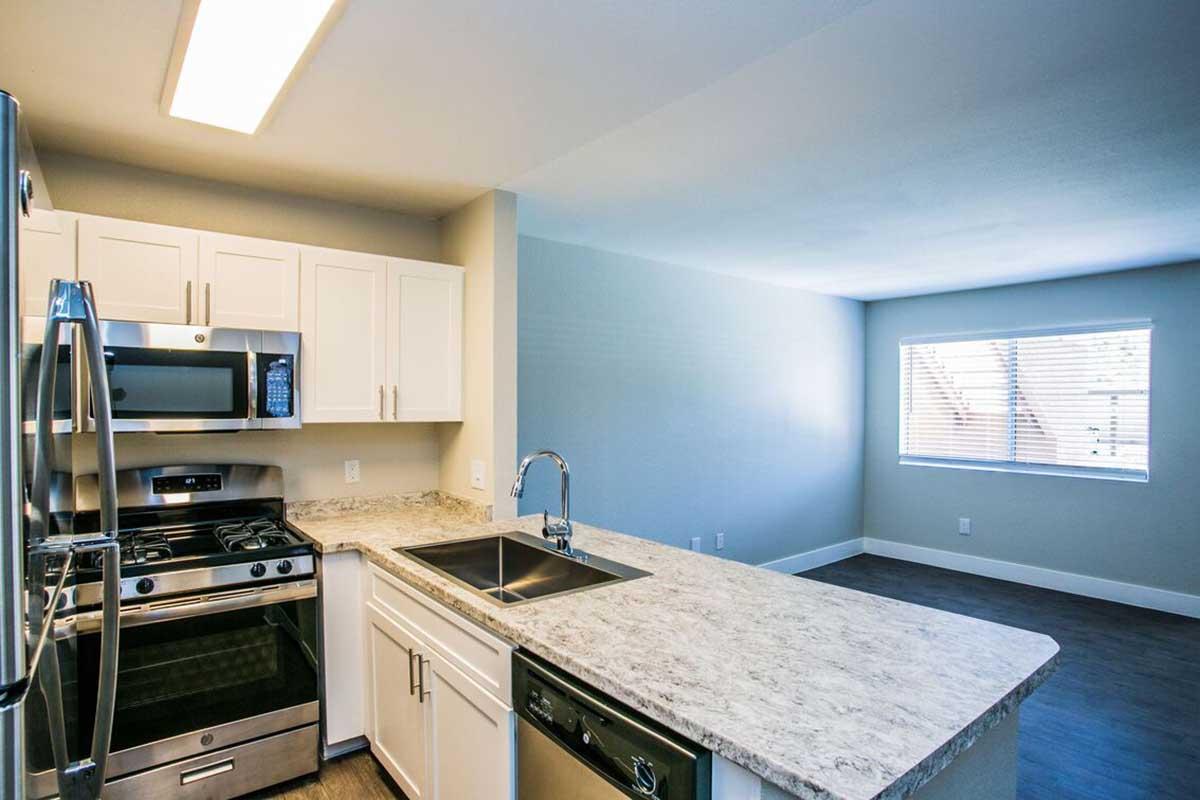 A modern kitchen featuring stainless steel appliances, including a gas stove, microwave, and a sink with a dishwasher. White cabinets complement the light granite countertop. The kitchen is open to a well-lit living area with a large window and neutral-colored walls.