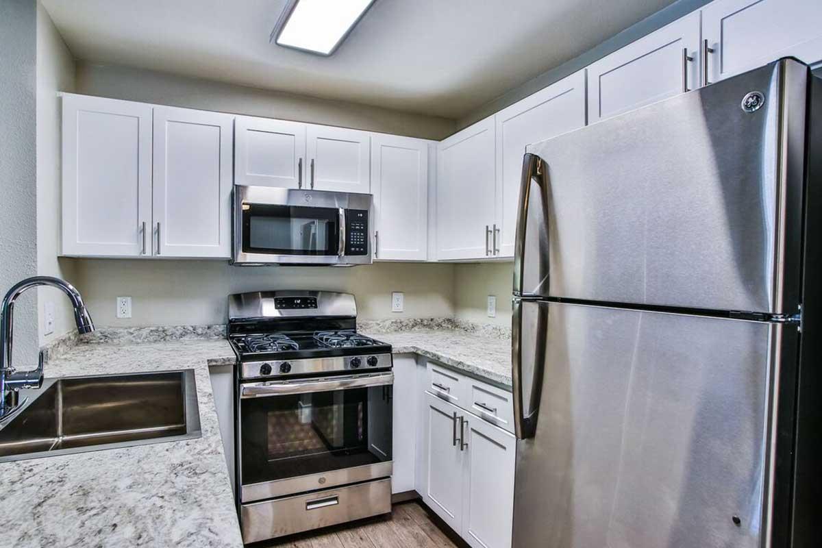 Modern kitchen featuring white cabinetry, stainless steel appliances including a microwave, a gas stove, and a refrigerator. The countertop is made of light-colored granite, and there is a sleek sink with a pull-down faucet. Bright overhead lighting enhances the spacious, well-organized design of the kitchen area.