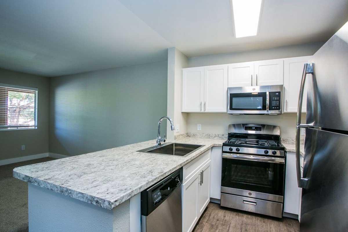 Modern kitchen featuring stainless steel appliances, including a gas stove, microwave, and dishwasher. The countertops are made of light granite, and the cabinetry is white. The space is well-lit with natural light coming from a window, showcasing an open layout with a serene color scheme.