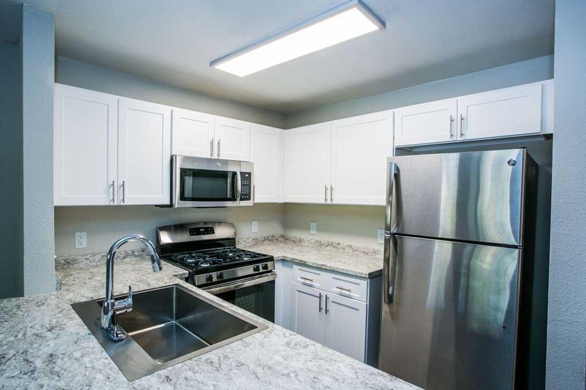 A modern kitchen featuring white cabinetry, stainless steel appliances including a microwave and refrigerator, a gas stove, and a sleek granite countertop with a sink. Natural light from a ceiling fixture brightens the space, enhancing the clean, contemporary design.
