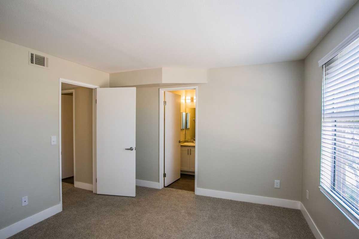 A modern, empty bedroom with light-colored walls and carpeting. Two doorways lead to adjacent areas, one of which has a bathroom visible. The room features a window with blinds that lets in natural light, creating a bright and spacious feel.