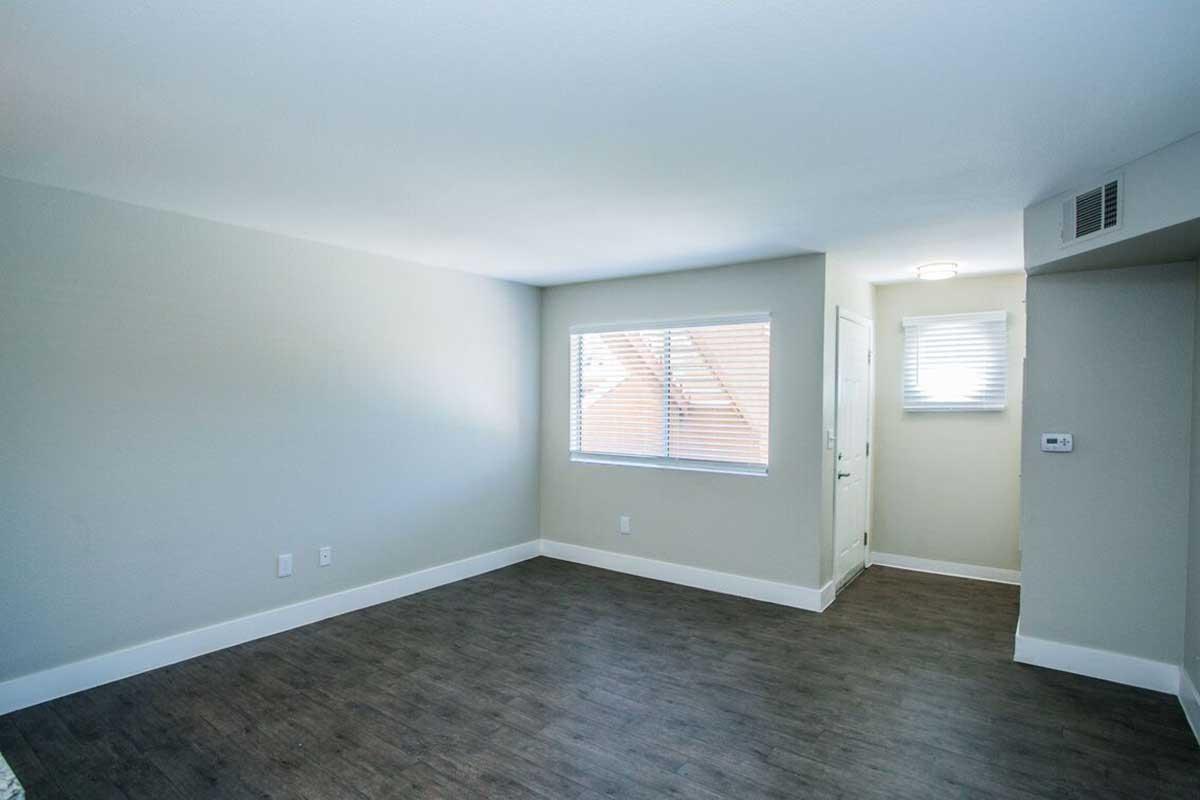 Empty interior of a room with light-colored walls and a window with blinds. The flooring is dark laminate. There is a door on one wall, and the space appears bright and well-lit.