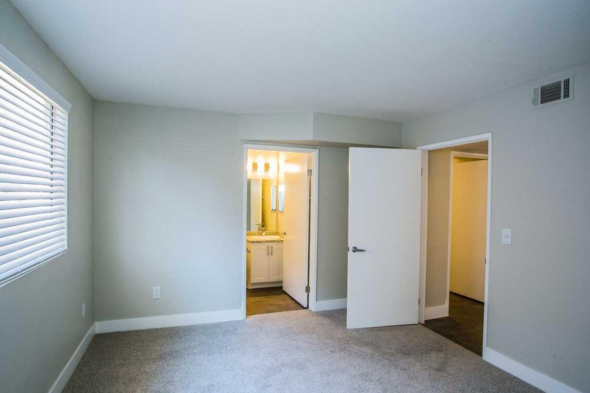 A spacious, light-colored bedroom with soft gray carpeting. The room features a window with horizontal blinds on the left side, and an open doorway leading to a bathroom area. Two additional doorways are visible, suggesting access to other rooms. The walls are painted a neutral tone.