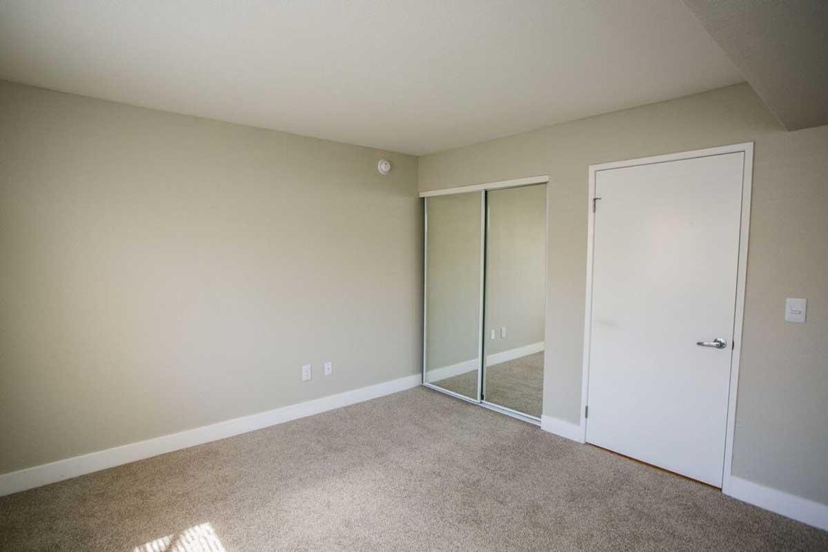 A well-lit, empty room with light beige walls and carpet. It features a large mirror closet along one wall and a simple white door. The space appears clean and spacious, providing a neutral backdrop suitable for various furnishings.