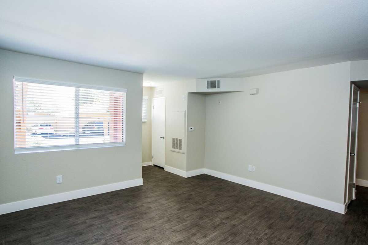 A spacious, empty living room with light-colored walls and dark wood laminate flooring. There are horizontal blinds on the window allowing natural light in, and a small closet is visible on the right side. The overall atmosphere is bright and modern, suitable for various interior design styles.