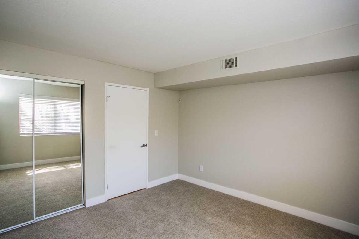 Empty room with light-colored walls and carpeted floor. It features a closet with mirrored sliding doors and a white door. Natural light enters through a window covered with blinds, creating a bright and spacious ambiance. The room is unfurnished, giving it a clean and minimalist appearance.