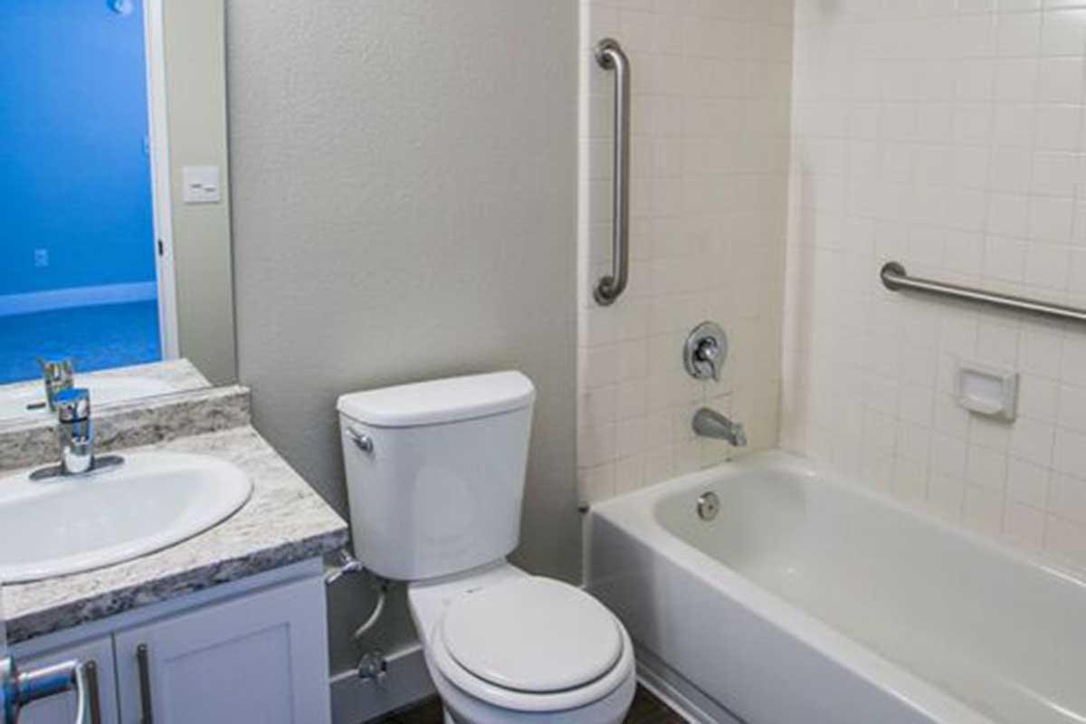 A clean and modern bathroom featuring a white bathtub with a showerhead, a toilet, and a granite countertop sink. The walls are painted in a neutral color, and there is a grab bar next to the bathtub for safety. A door leads to another room, adding a touch of spaciousness.