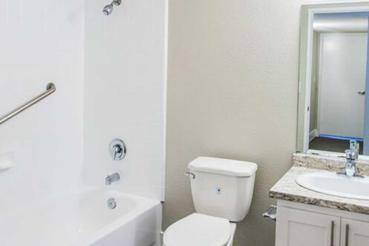 Bright, modern bathroom featuring a bathtub with showerhead, a white toilet, and a granite countertop with a sink. The walls are painted a soft color, and there is a mirror above the sink. A doorway in the background leads to another room, indicating an open layout.