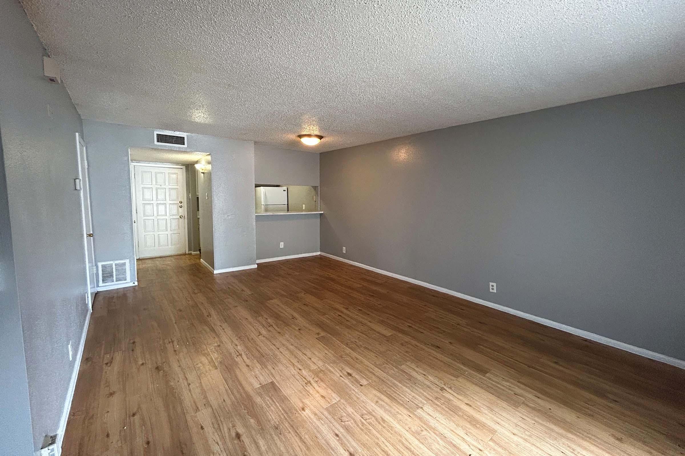 A spacious living room with gray walls and laminate flooring. There is a door leading to another room, and a small kitchenette area visible in the background. The space is well-lit, featuring a ceiling light and a neutral color scheme, creating a simple and inviting atmosphere.