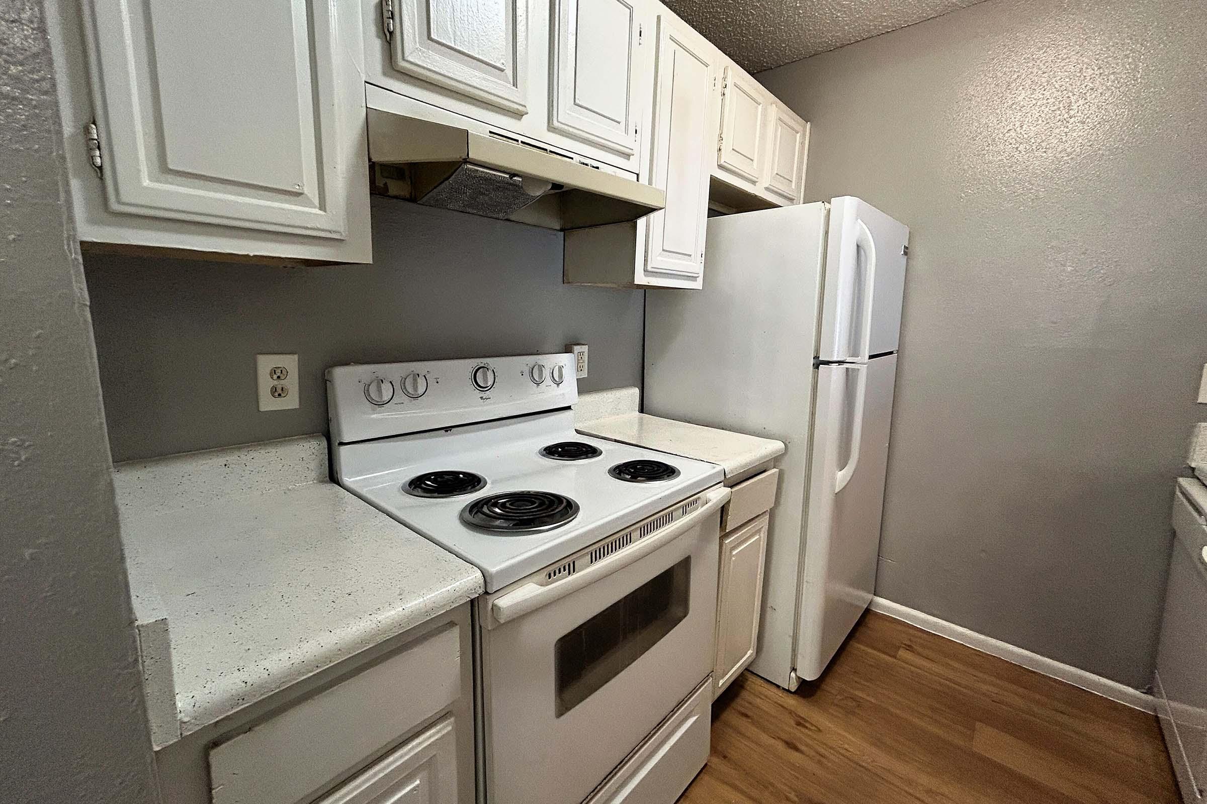 A compact kitchen featuring white cabinets, a white stove with an oven, a range hood, and a white refrigerator. The countertop is light-colored, and the flooring is laminate wood. The walls are painted in a light gray color, creating a clean and simple aesthetic.