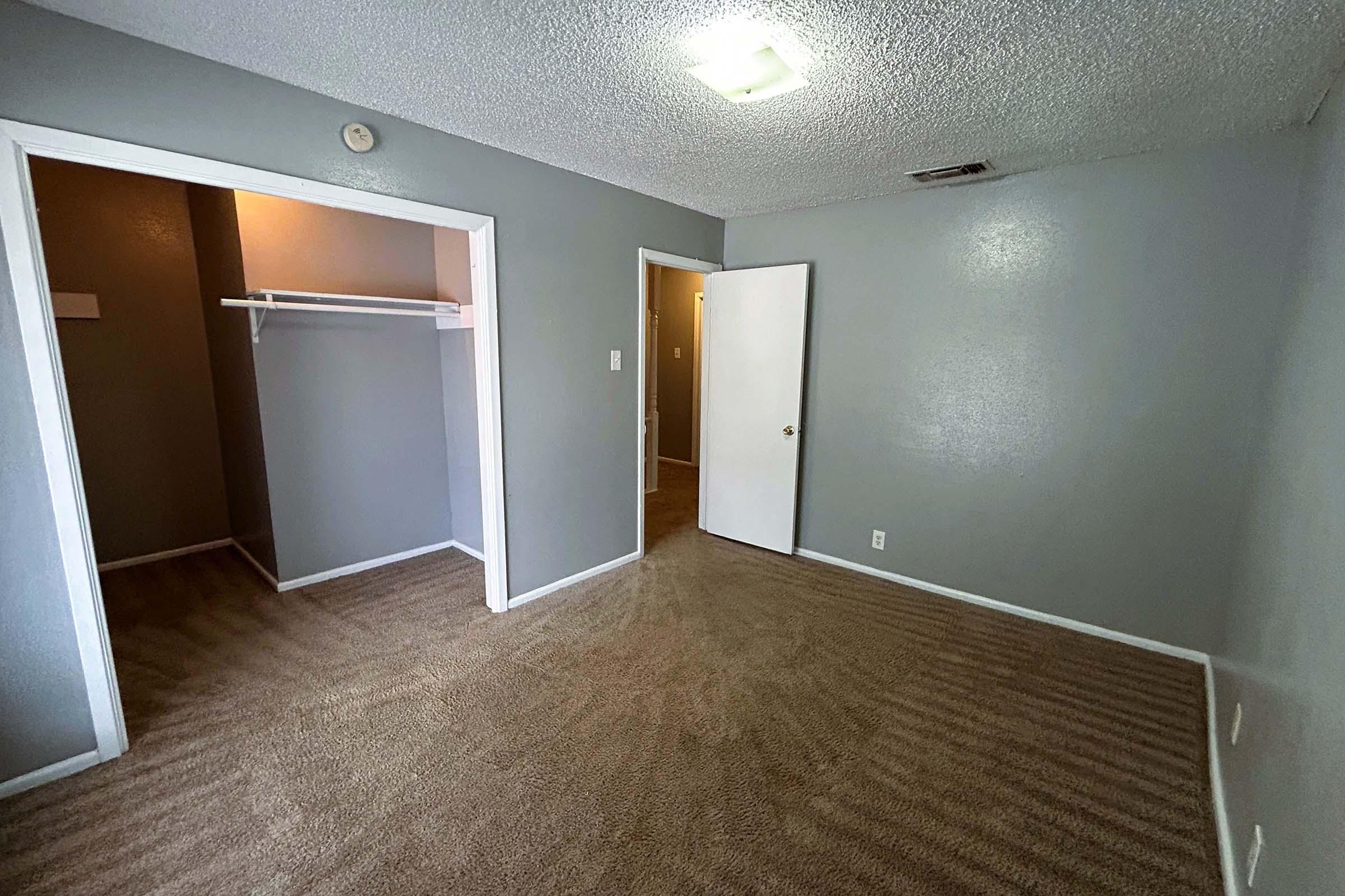 A spacious, empty room with gray walls and carpeted flooring. There is a small closet on the left with a hanging rod, a door leading to another room, and a light fixture on the ceiling. The overall ambiance is bright and neutral, suitable for various decorating options.