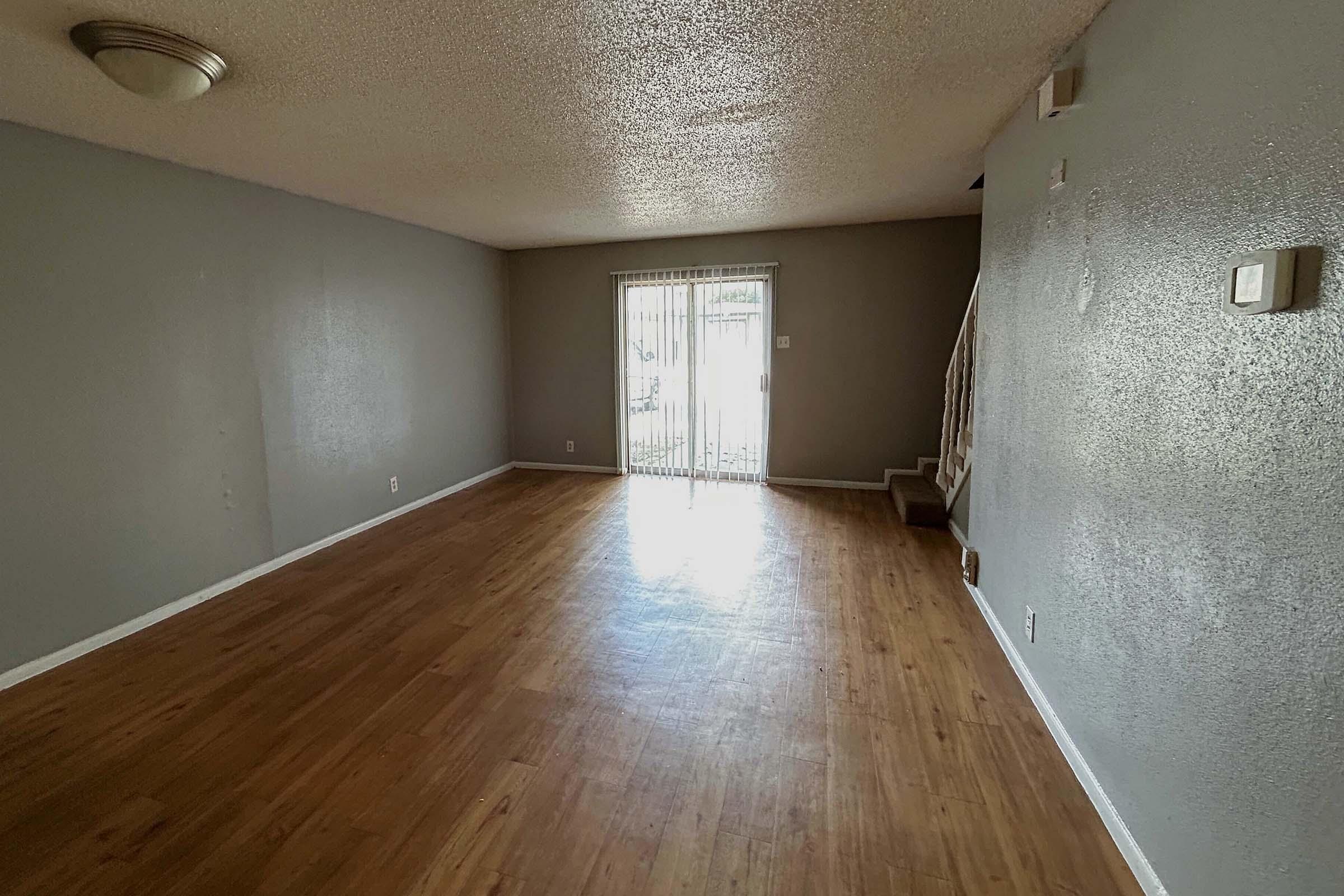 An empty living room with wooden flooring, gray walls, and a sliding glass door leading outside. A staircase is visible in the background, and a ceiling light fixture is installed. The space is bright and airy, with natural light coming through the door.