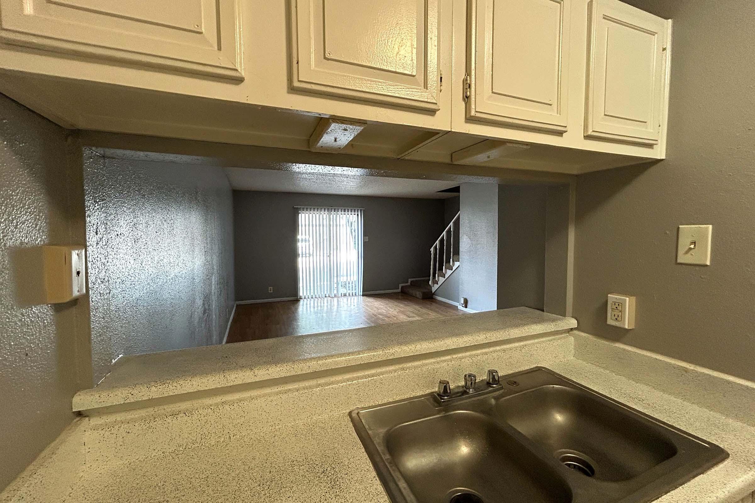 A view from a kitchen with a double sink, looking into an open living area. The living space features hardwood flooring and a sliding glass door with vertical blinds, leading to a staircase in the background. The walls are painted light gray, giving a modern feel.