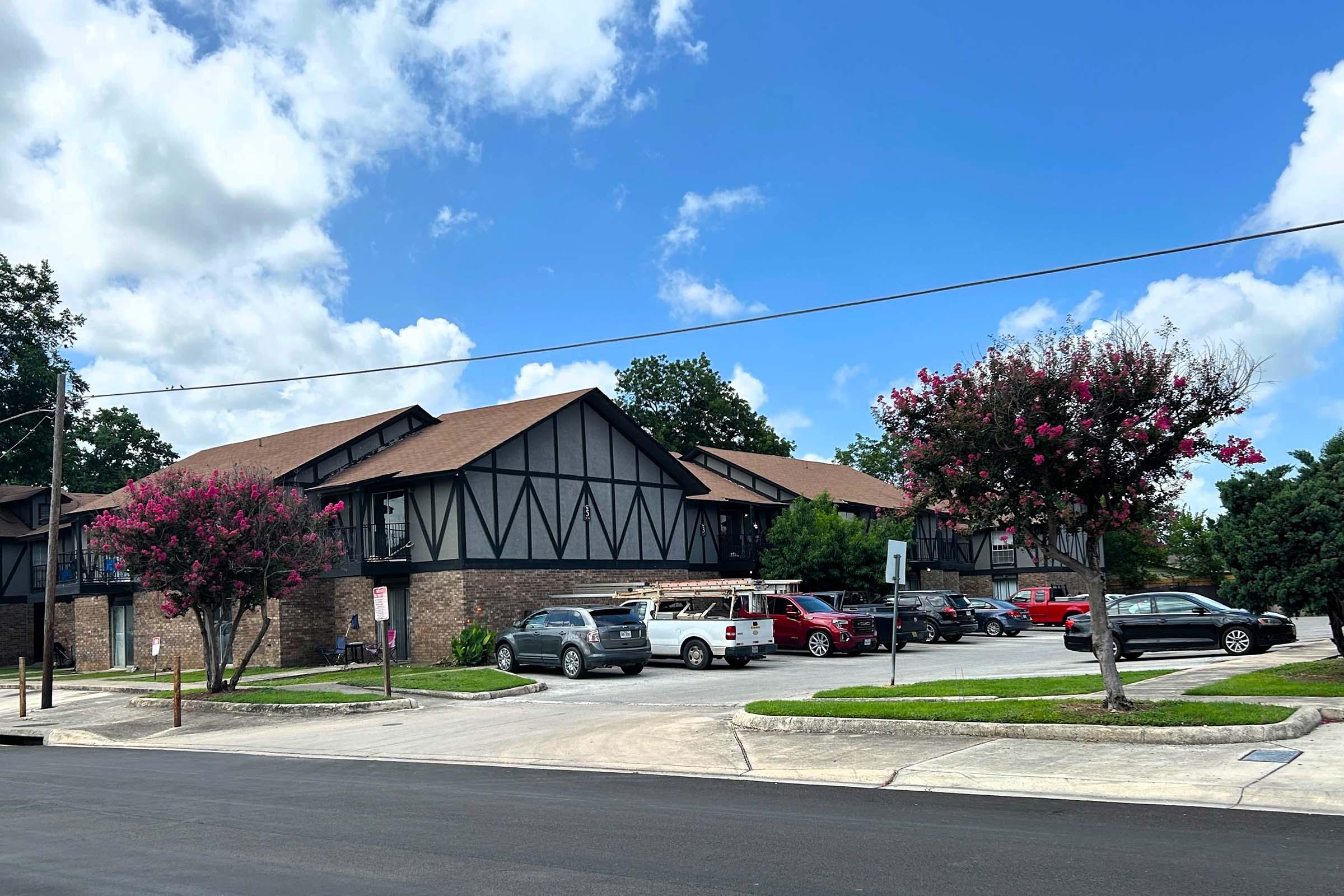 A view of a residential building featuring Tudor-style architecture, surrounded by green trees and blooming crepe myrtle flowers. In the parking area, several cars are parked, including red and gray vehicles. The sky is partly cloudy, creating a bright atmosphere.