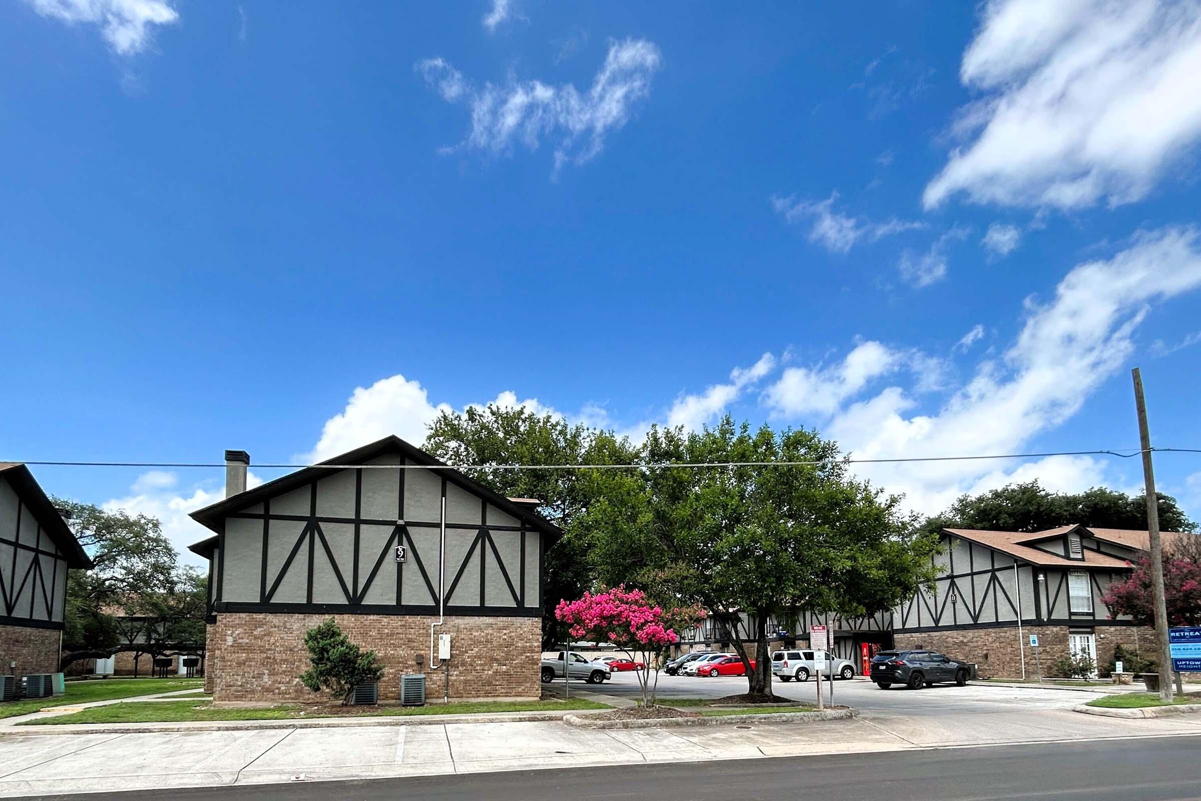 A row of two-story buildings designed in a Tudor style, characterized by dark wood accents and white stucco. A vibrant pink flowering tree is in front, along with parked cars. The sky is bright blue with fluffy white clouds, creating a sunny, pleasant atmosphere in a residential area.