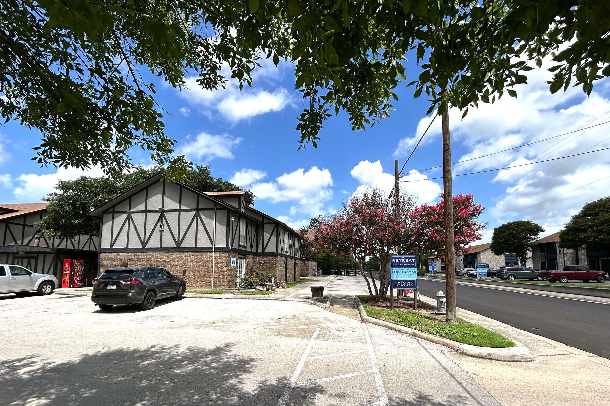 A parking lot with a building featuring a Tudor-style design. Green trees and pink flowering plants are visible in the foreground. The sky is partly cloudy with blue patches, and there are a few cars parked along the street in the background. A sign is also present near the building entrance.