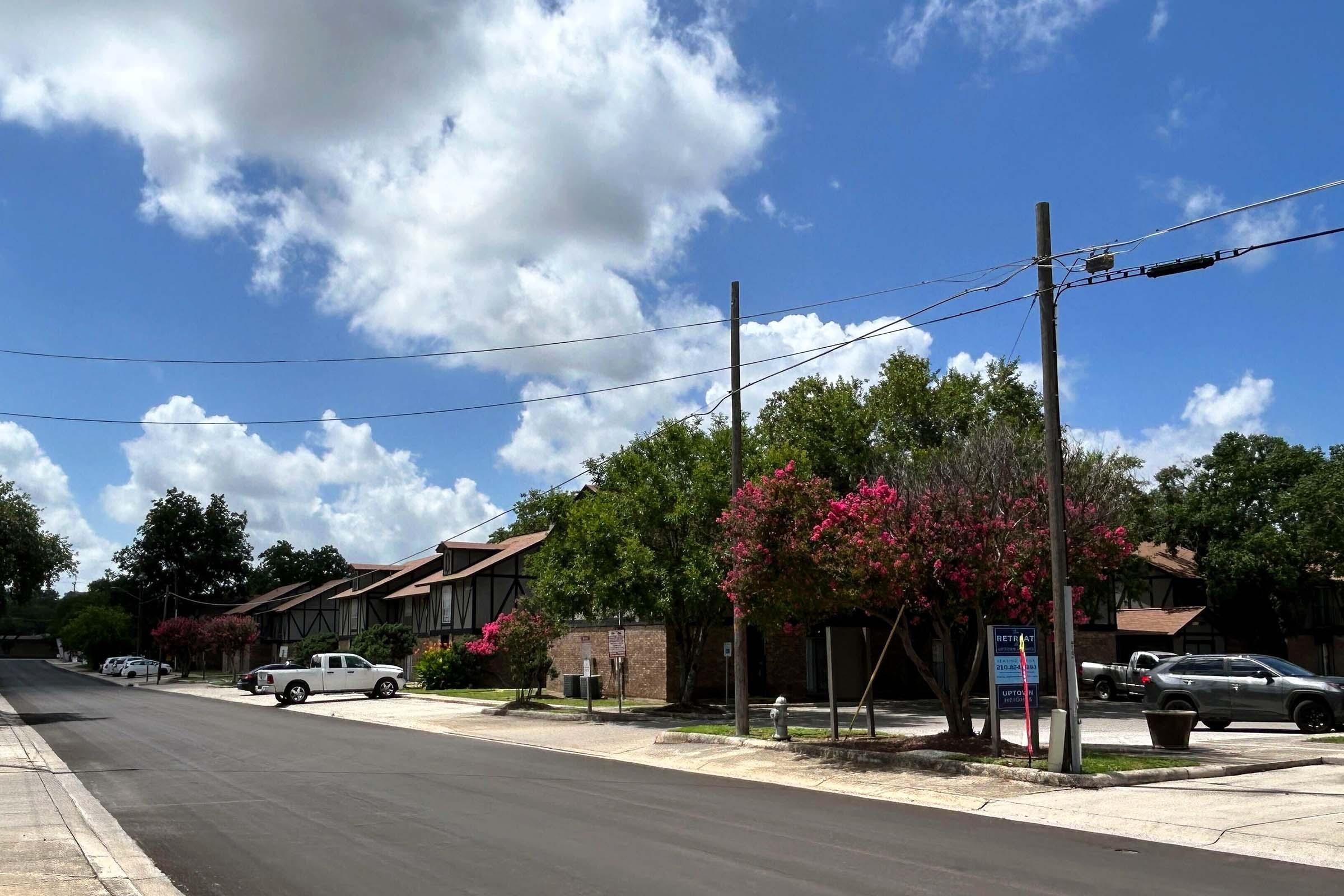A street view featuring two-story buildings with a brown and beige facade, surrounded by green trees and blooming pink crepe myrtle flowers. The sky is partly cloudy with blue and white clouds. Cars are parked along the side of the road, and there are utility poles along the street.