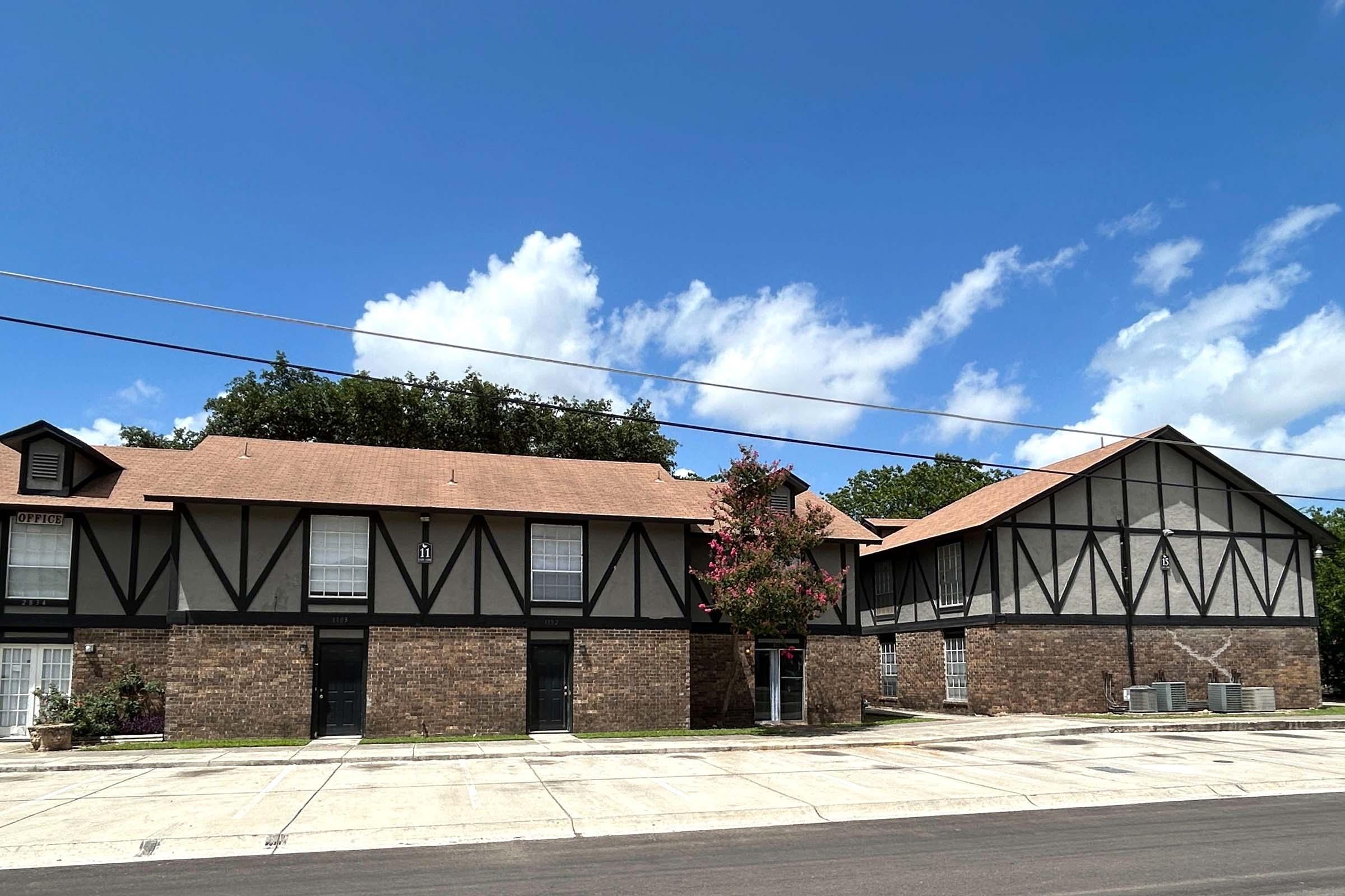 Two-story buildings with a mix of brick and Tudor-style architecture. The left building features dark wooden beams and white walls, while the right has a more plain design. A green tree with pink flowers stands in front, and a clear blue sky with fluffy clouds is visible above.