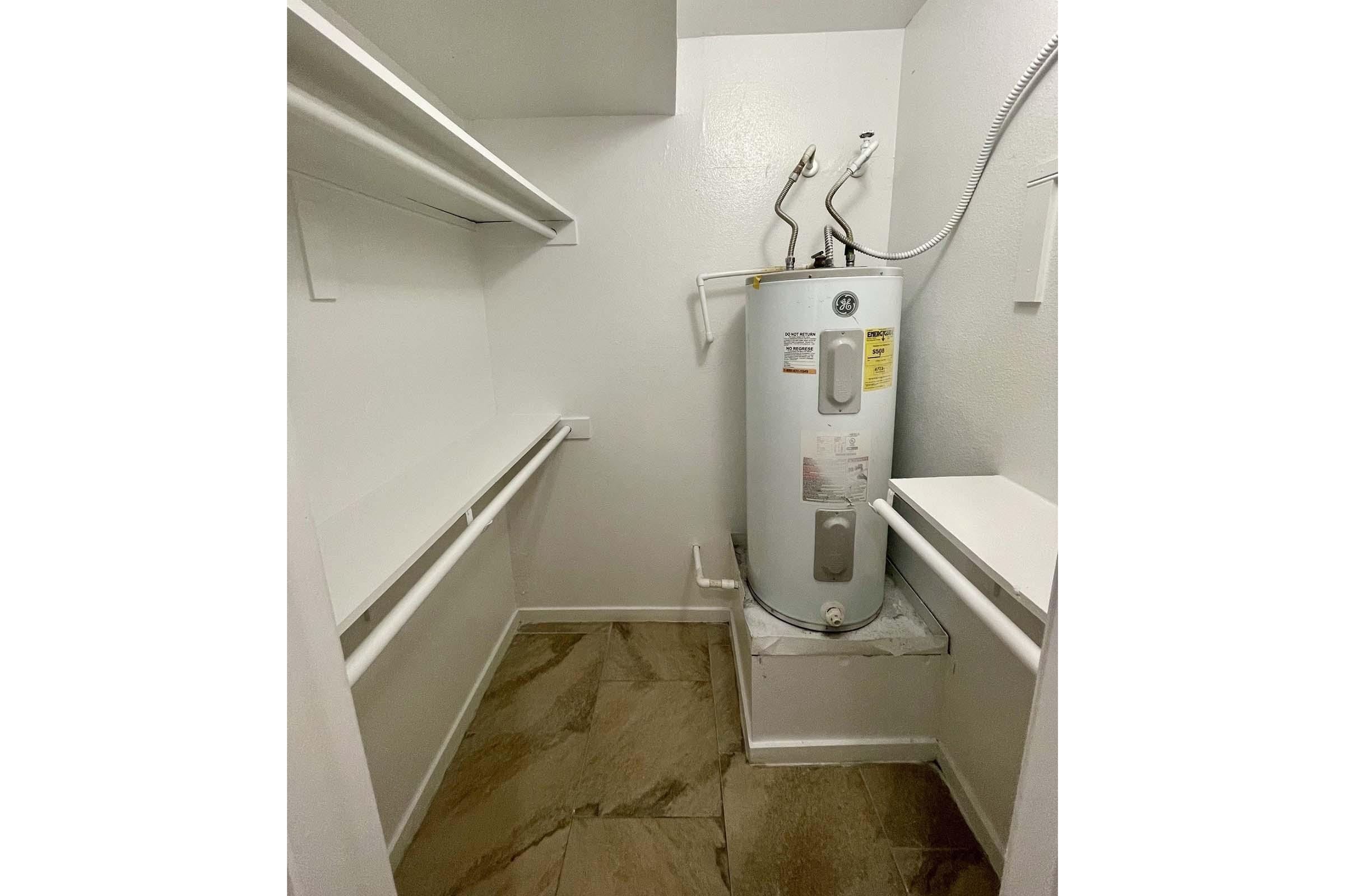 A small utility room featuring a white water heater against the wall, with shelves above and a countertop on the side. The floor is tiled with a light brown pattern, and the walls are painted white, creating a clean and minimalist appearance. The space is well-lit and organized.