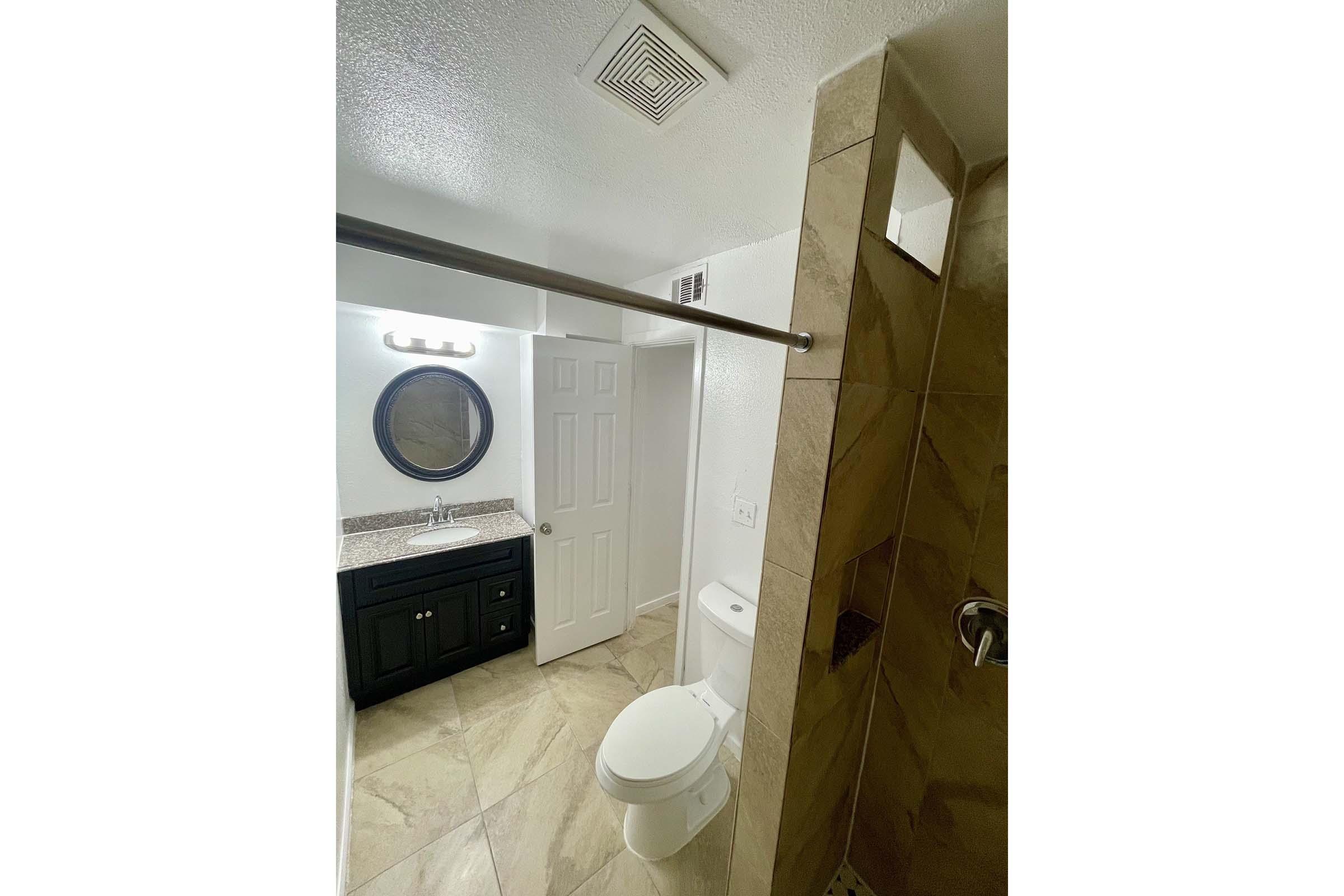 A modern bathroom featuring a shower, toilet, and vanity with a round mirror. The space has neutral-colored tile flooring, light walls, and a granite countertop. A towel bar is installed above the toilet, and there's an exhaust fan in the ceiling.