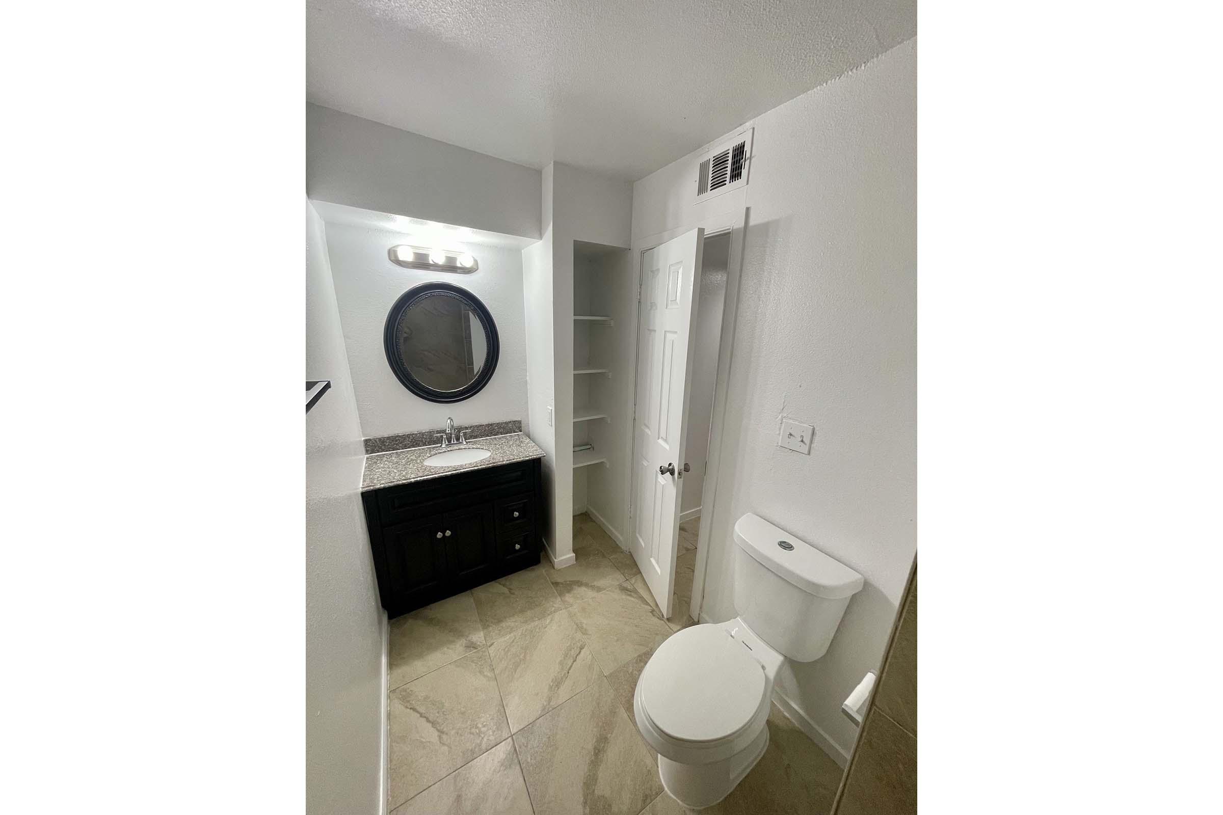 A modern bathroom featuring a round mirror above a granite countertop with a dark cabinet underneath. To the right, there is a white toilet, and to the left, a built-in shelving unit. The walls are painted white, and the floor has large light-colored tiles, creating a clean and bright appearance.