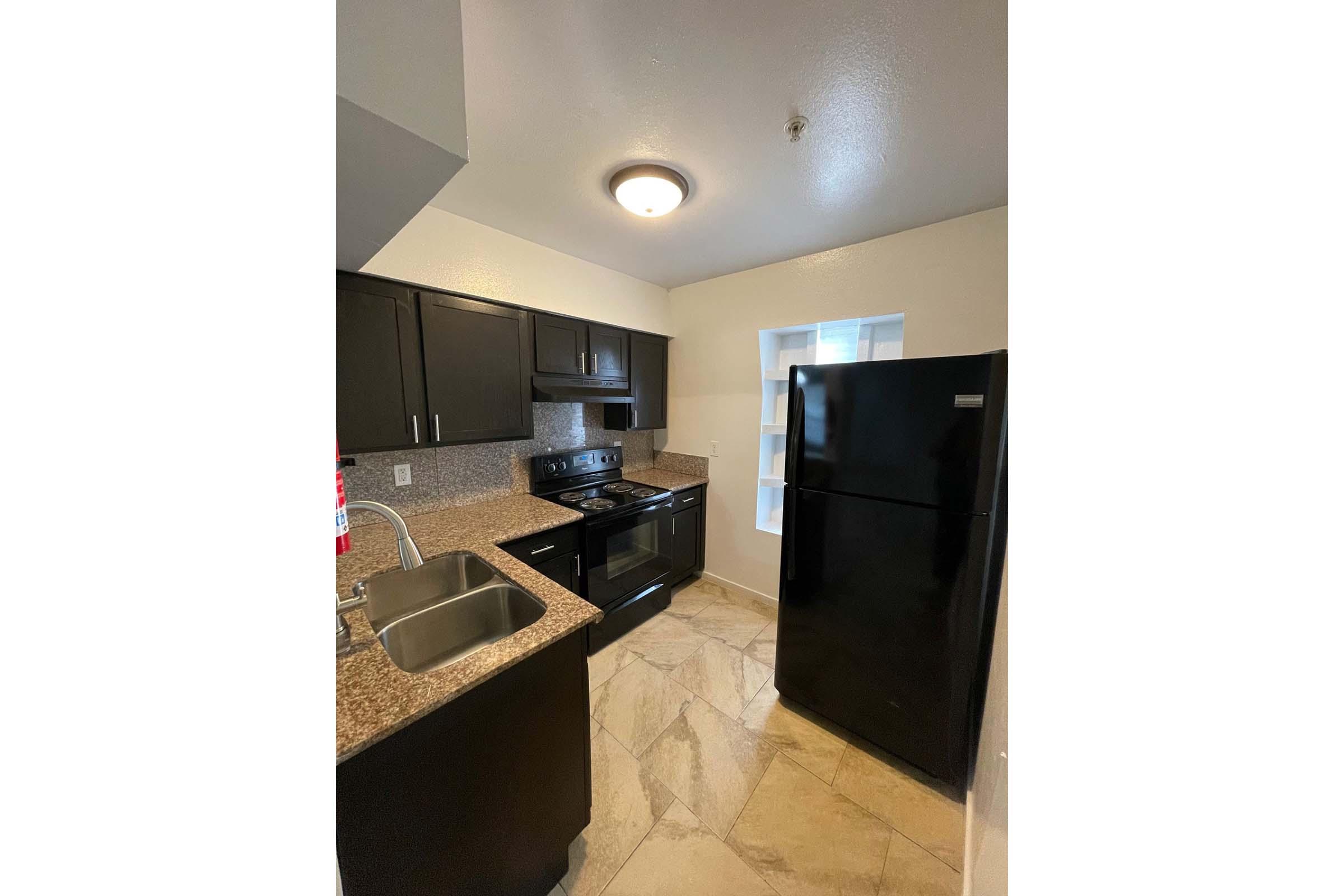 A modern kitchen featuring dark cabinetry, a black refrigerator, and a gas stove. The countertops are speckled granite, and the space has light-colored walls. There's a window providing natural light, and the floor is finished with light-colored tiles.