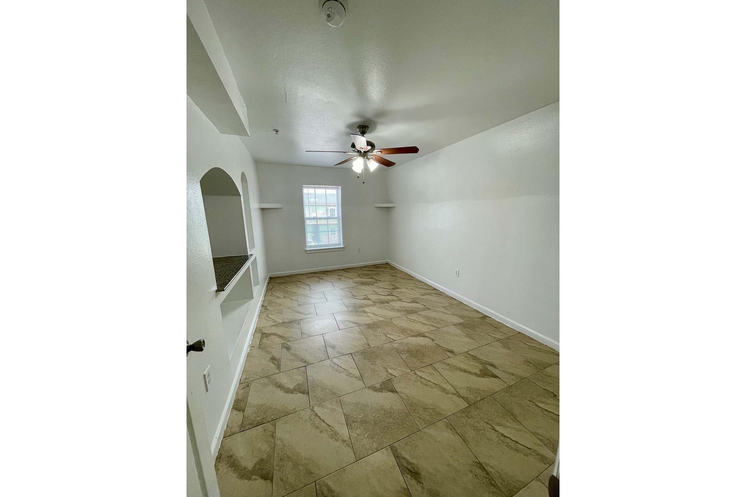 A bright, empty room featuring light-colored walls, a ceiling fan with wooden blades, and tiled flooring. There are two wall shelves and a window allowing natural light to enter the space. The room is designed for potential furniture arrangement and personalization.