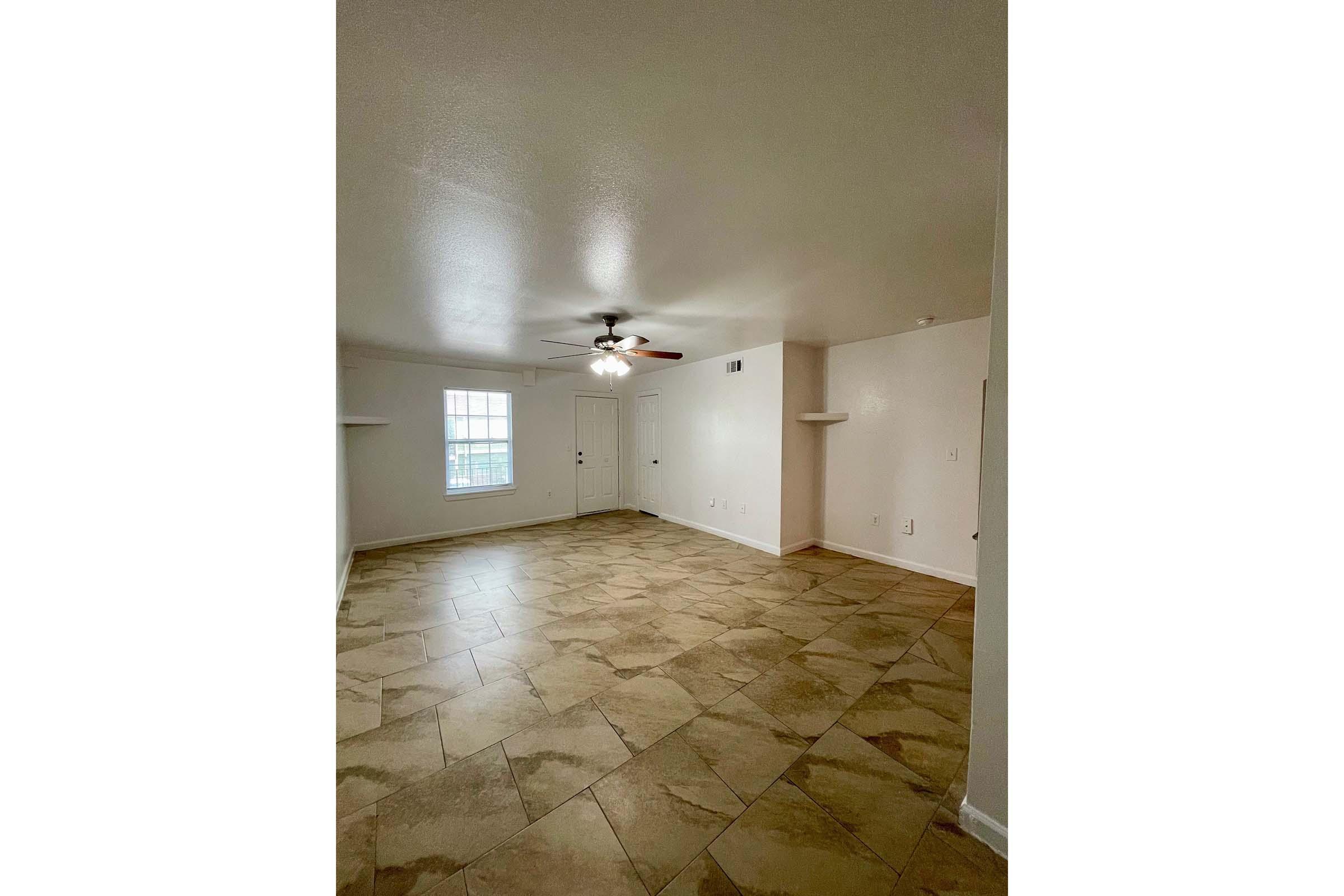 A spacious, empty room with a ceiling fan and light fixture. The walls are painted white, and the floor features large, light-colored tiles. A window lets in natural light, and there's a door visible, leading outside. There are small shelves on either side of the room.
