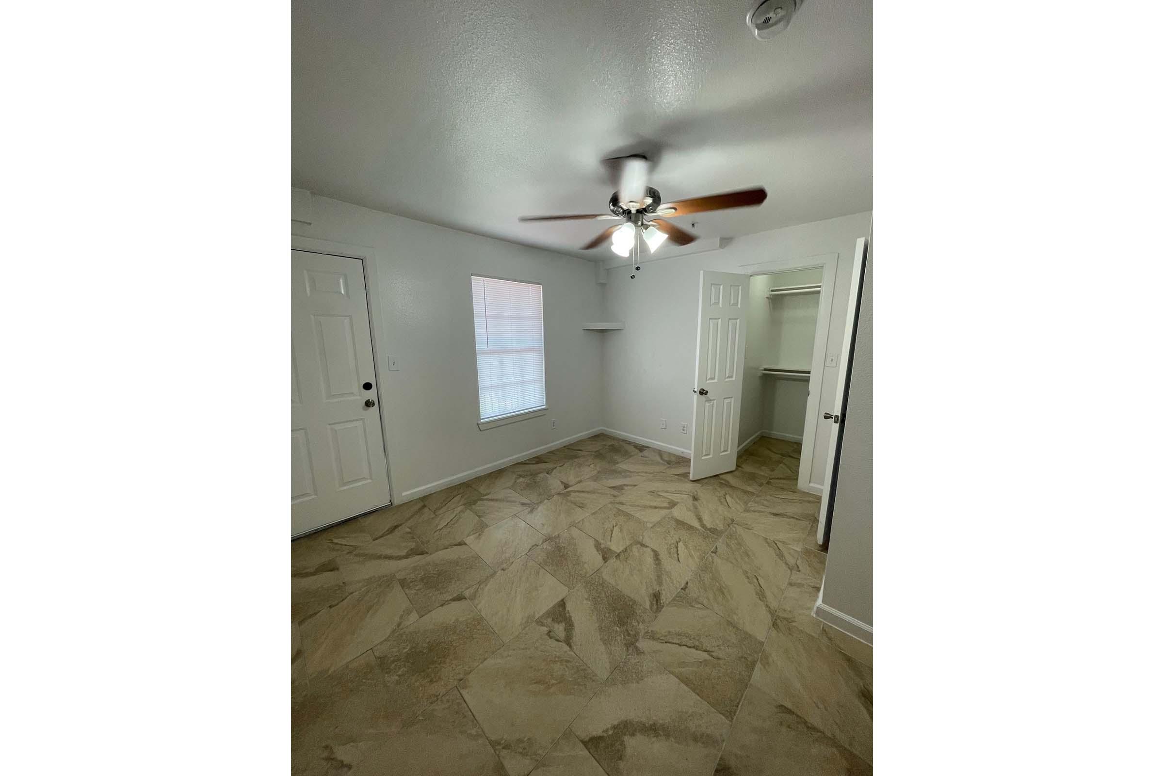 A well-lit room with neutral gray walls and a ceiling fan. It features two white doors: one leading outside and the other to a closet. A window with white blinds allows natural light in, illuminating the patterned floor tiles. The space is clean and ready for decoration.