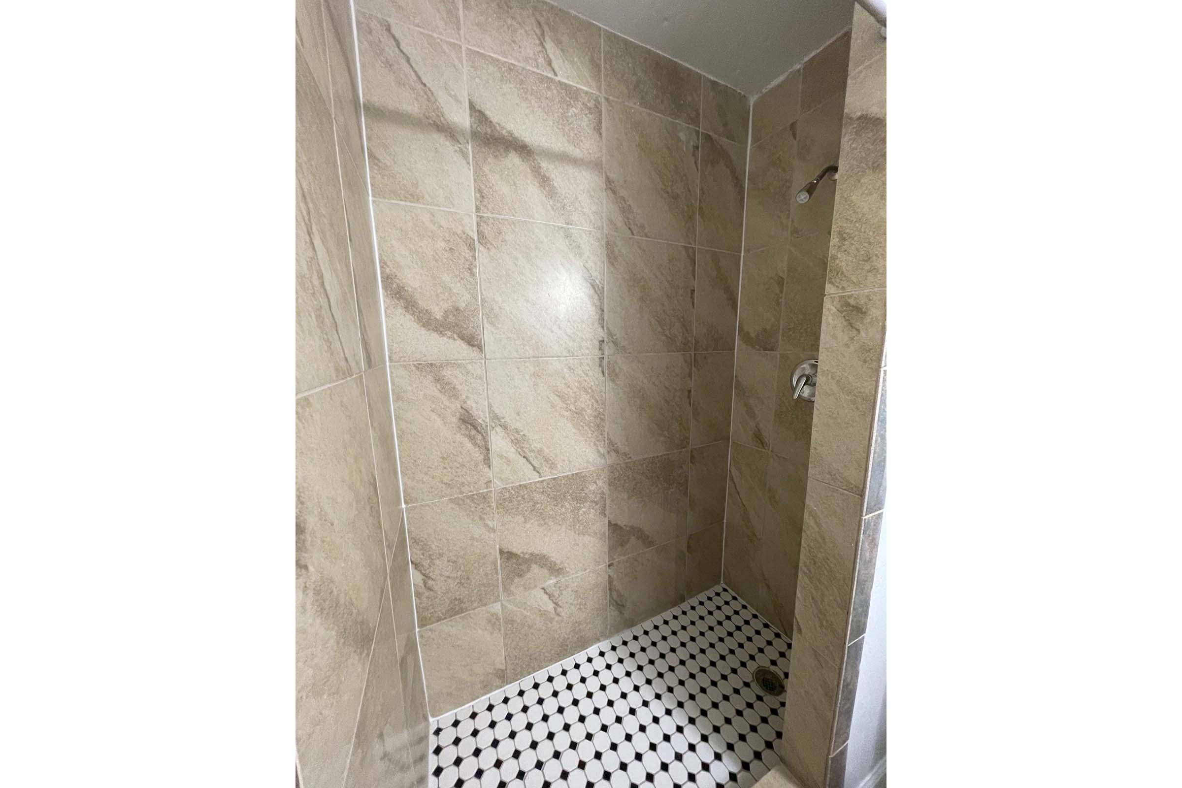 A tiled shower space featuring beige and brown marble-like wall tiles, with a black and white patterned floor. There are two showerheads, one mounted on the wall and another handheld, creating a modern and clean design. The space is well-lit, showcasing the tile textures.