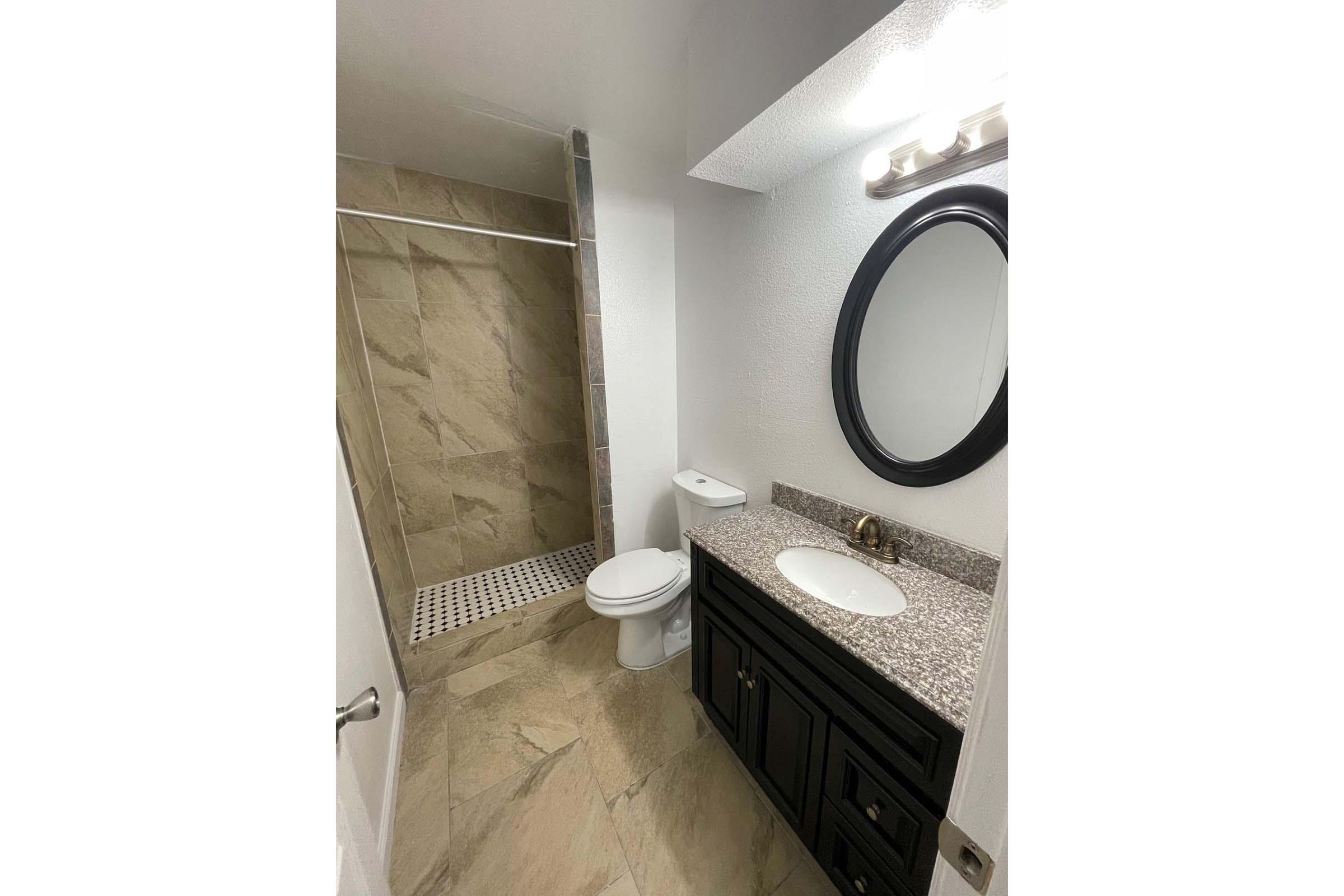 A modern bathroom featuring a shower with glass doors, a toilet, and a vanity with a round mirror and sink. The floor is tiled with large beige tiles, and the walls are painted white. The space is well-lit and clean, with a neutral color palette.