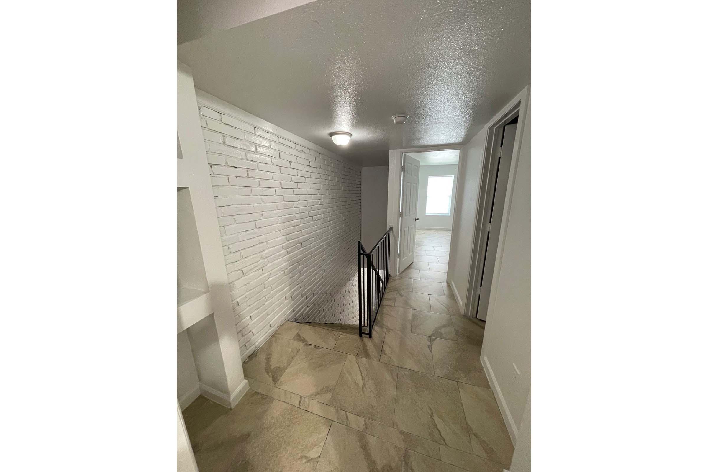 A well-lit interior hallway with a white brick wall on one side and a staircase leading down. The floor is tiled in a light color, and doors are visible at the end of the hallway. The space appears modern and clean, with neutral colors and a bright ambiance.