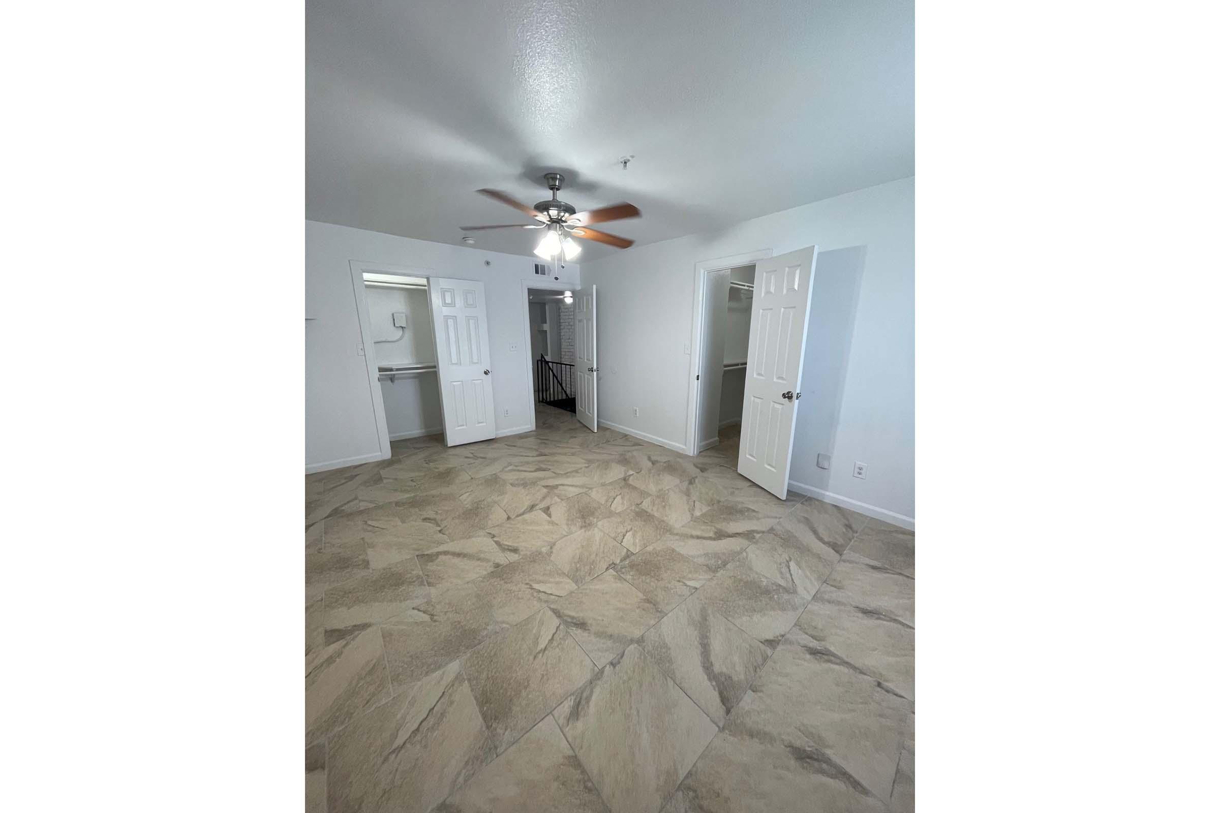 A spacious, well-lit room featuring light-colored walls and a ceiling fan. The floor is covered in large, patterned tiles. There are two white double doors leading to separate areas, with a staircase visible in the background. The room has an open layout, creating a welcoming atmosphere.