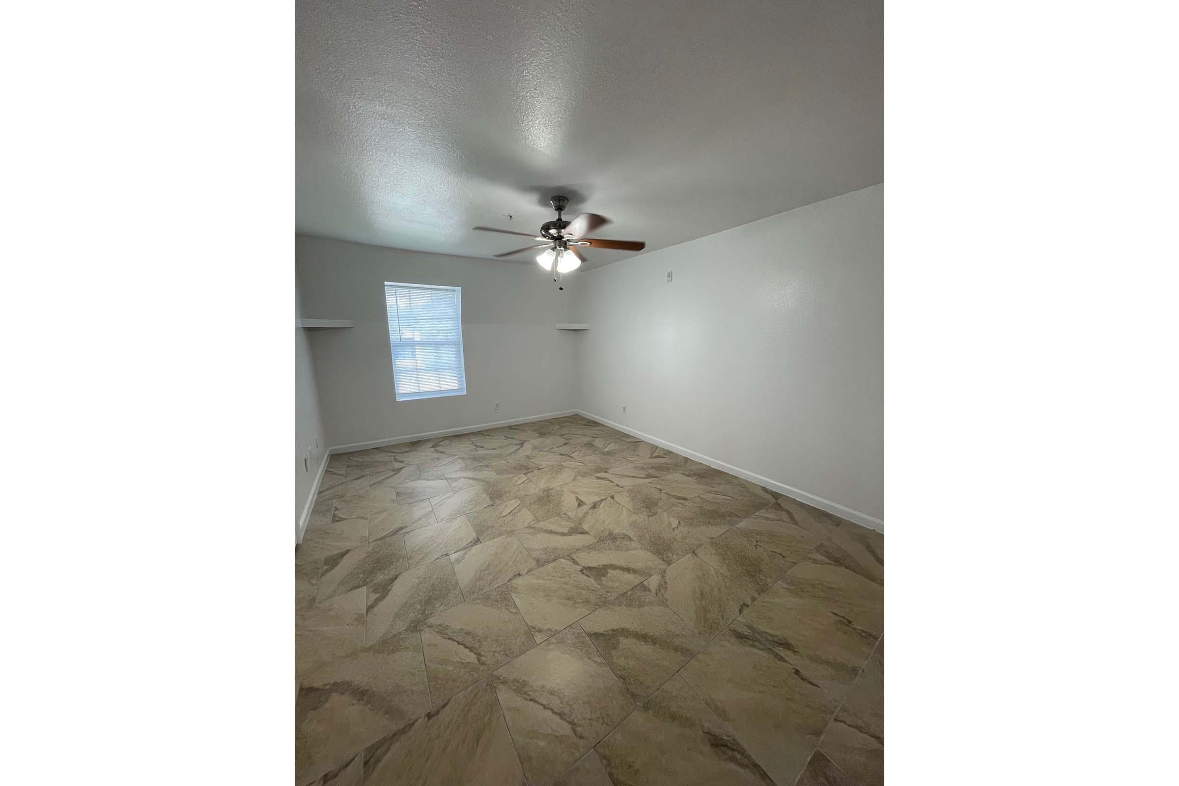 A spacious, empty room with light-colored walls and a ceiling fan. The floor is covered with large, patterned tiles. A single window lets in natural light, casting a soft glow on the surroundings. The room is unfurnished, creating an open and airy atmosphere.