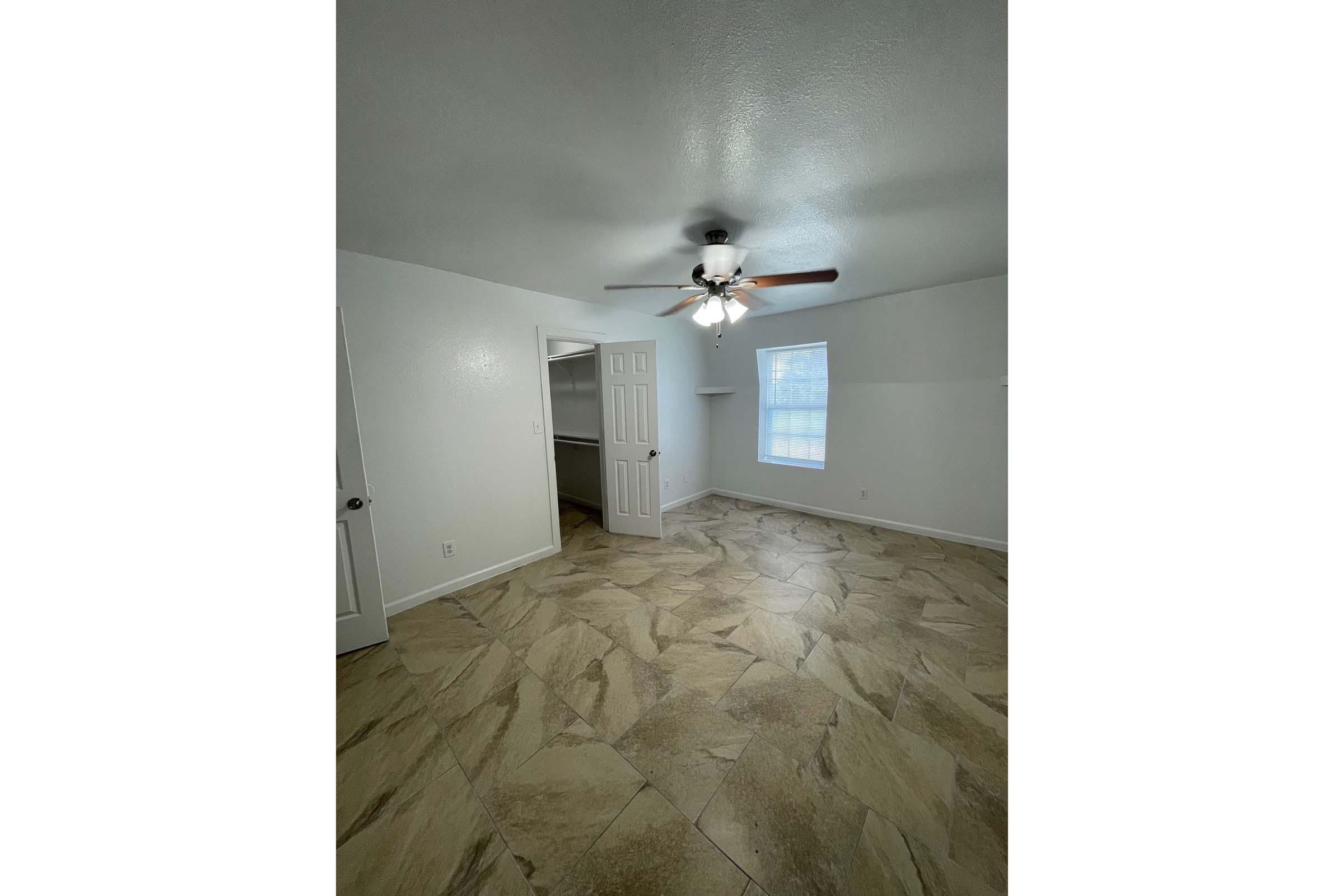 A spacious, well-lit room featuring light-colored walls and a ceiling fan. The floor is tiled with a marble-like design. A closet door is visible on the left, and a window lets in natural light. The room has a clean and open feel, suitable for various uses like a bedroom or office.