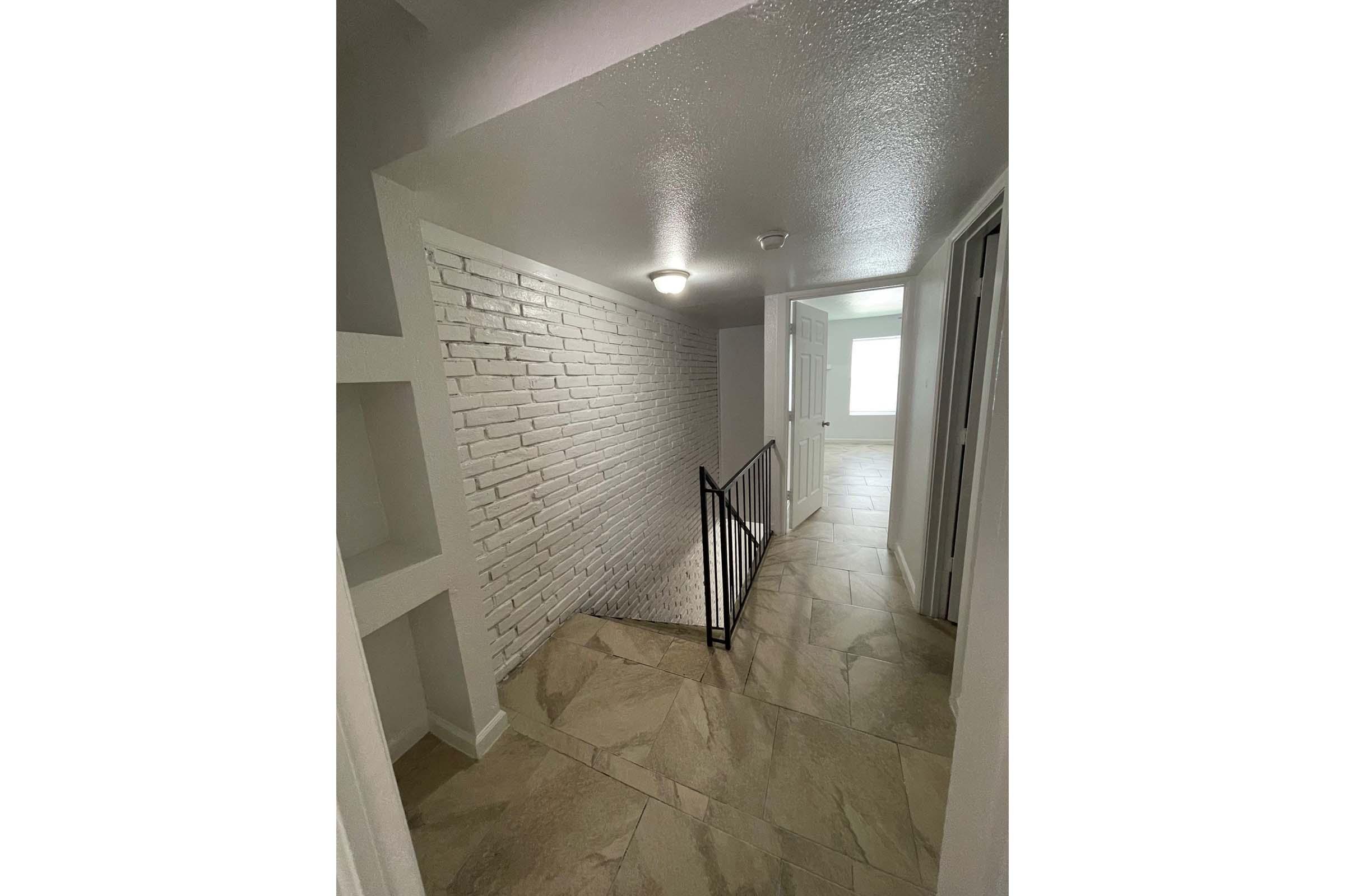 A well-lit hallway featuring a white brick wall and a staircase with a black railing. The floor is tiled in a light color, and there are doors leading to adjacent rooms. Natural light is entering from a window at the end of the hallway, creating an inviting atmosphere.
