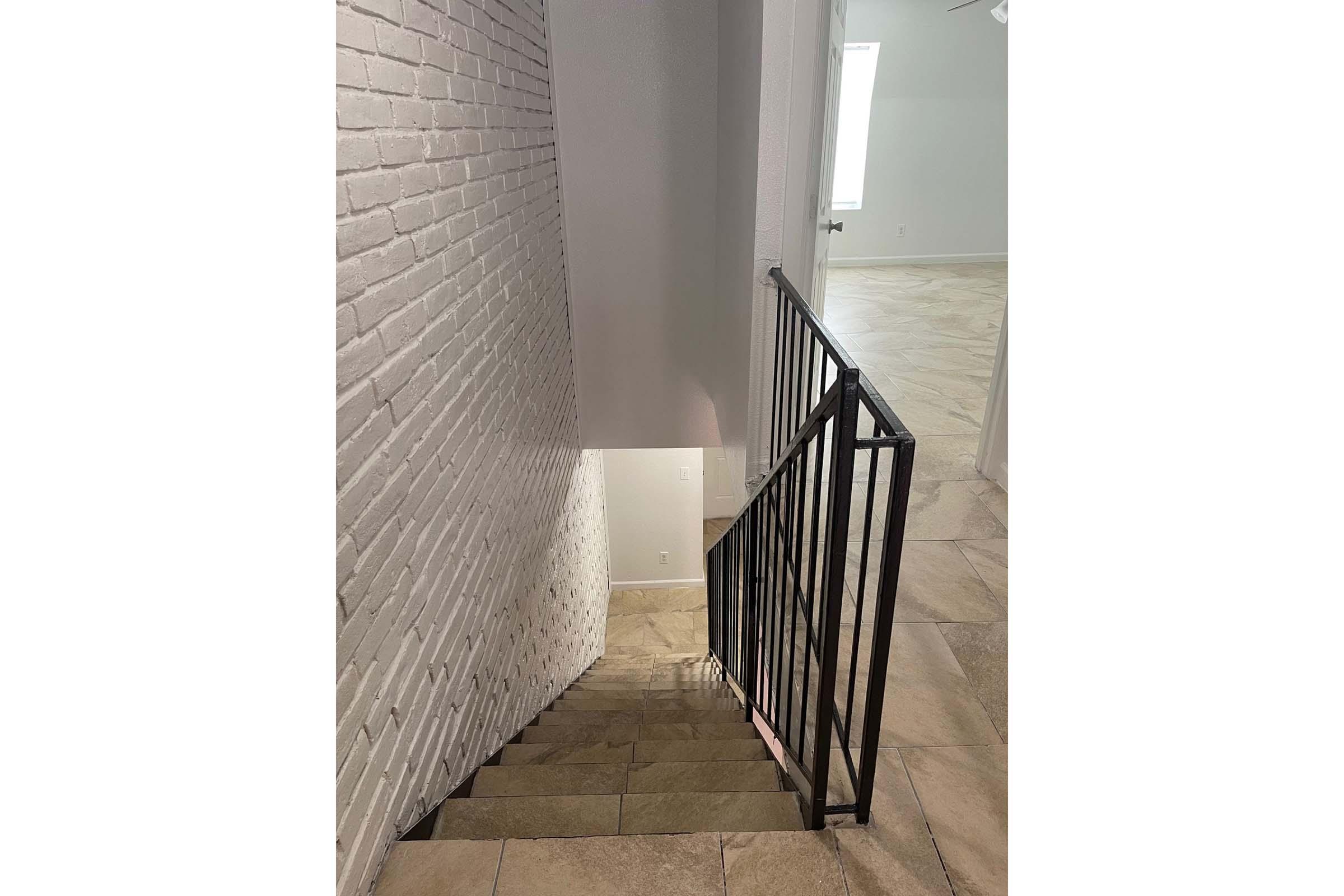 Interior view of a staircase leading downward, featuring a white brick wall on the left and black iron railing. The staircase has light-colored tiles, and the space below is illuminated by natural light from a window. The surrounding area appears to be a modern interior design.