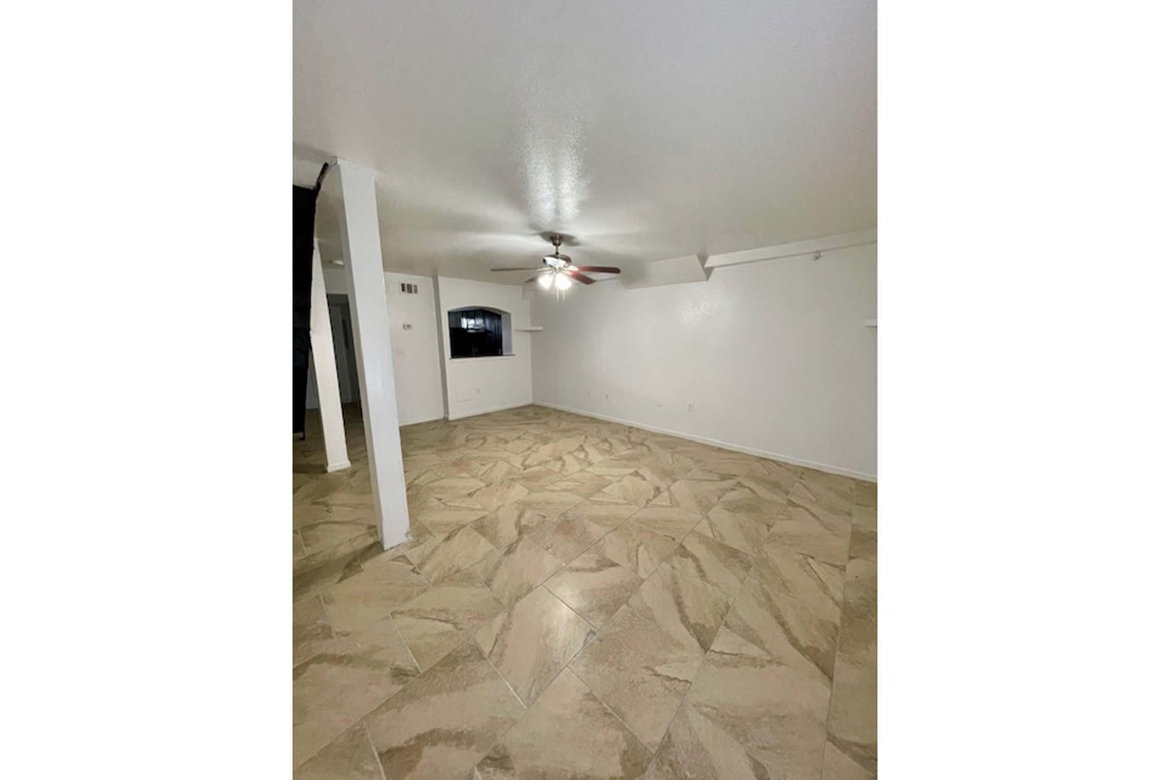 A spacious, empty room with beige tile flooring, a ceiling fan, and white walls. There is a wall-mounted shelf and a large window on one side. The room appears well-lit and ready for furnishing.
