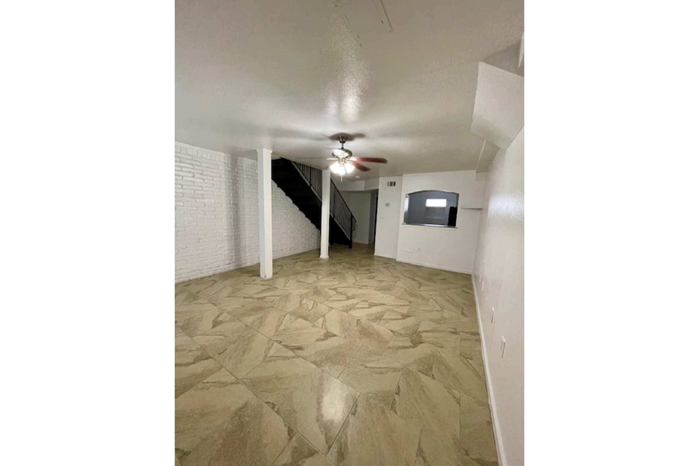 A spacious, empty room featuring light-colored tiled flooring, white walls, and a ceiling fan. To the left, a staircase leads to a second level, and a small window sits on the far wall, allowing natural light into the space. The overall ambiance is bright and airy.