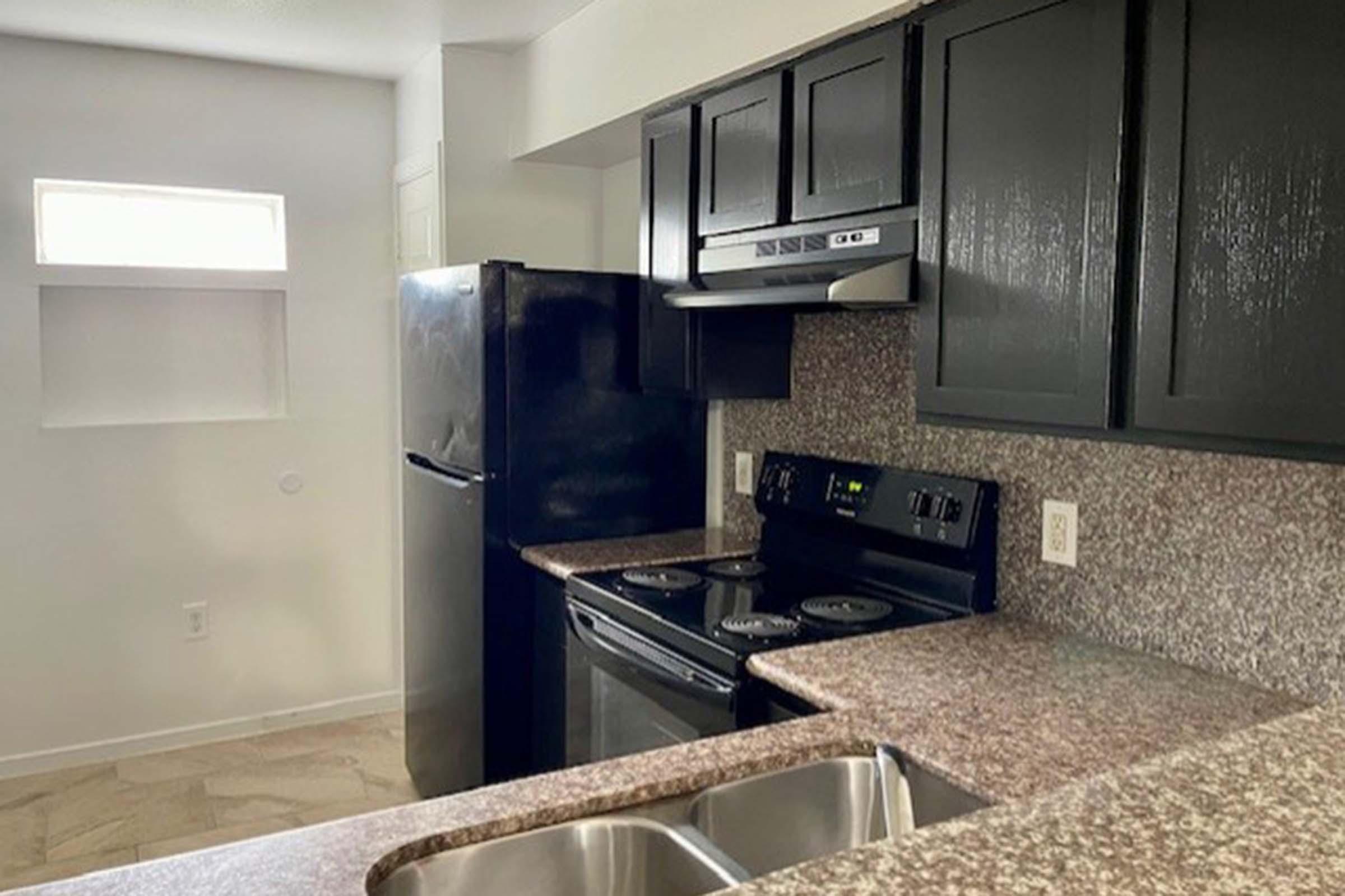 A modern kitchen featuring dark cabinetry, a black refrigerator, and a black electric stove. The countertop is made of speckled granite, and there are dual sinks. A small window above the sink allows natural light into the space. The floor consists of light-colored tiles.