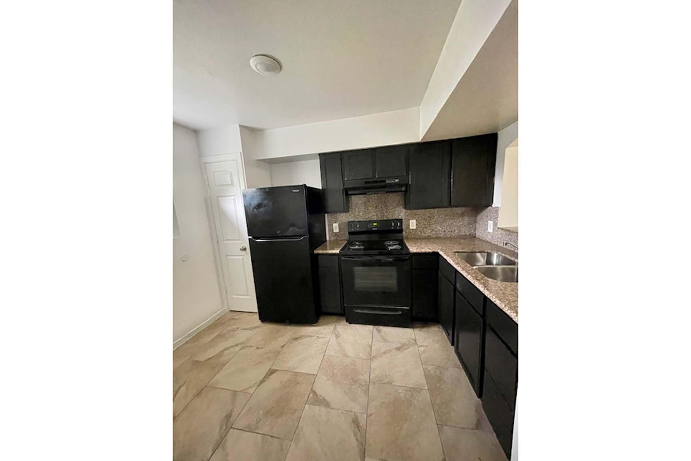 A modern kitchen featuring black cabinets and appliances, including a refrigerator and stove. The countertops are made of light-colored granite, and there is a sink next to a window. The flooring is tiled in a beige tone, creating a clean and contemporary look. The space is well-lit and organized.