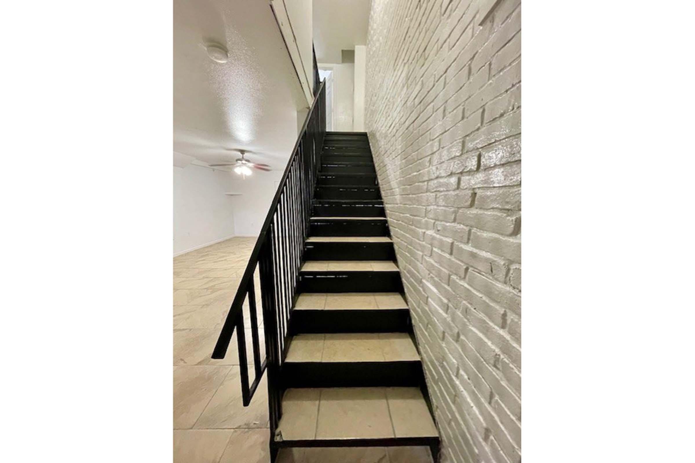 A set of black metal stairs leading upward, against a white brick wall. The stairs are well-lit and the surrounding area features a light-colored tiled floor. The space above is empty and open, with a ceiling fan visible to the left.