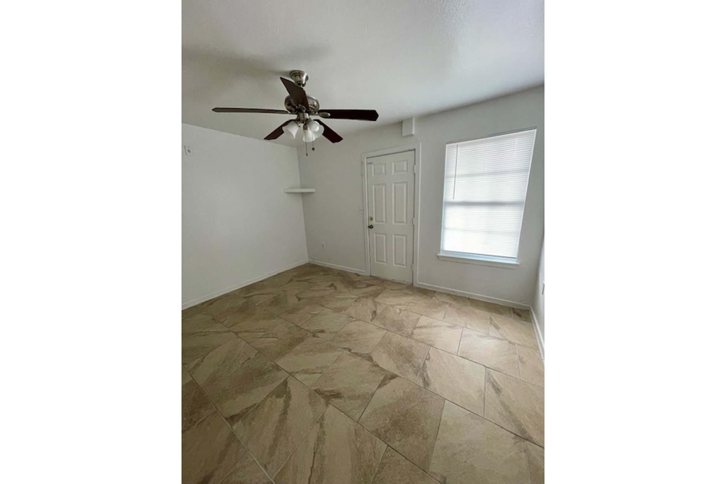 A spacious room with light-colored tiled flooring, featuring a ceiling fan. There is a white door leading outside and a window with blinds that lets in natural light. The walls are painted in a light shade, and there's a small shelf on one side. The room appears clean and empty, ideal for various uses.