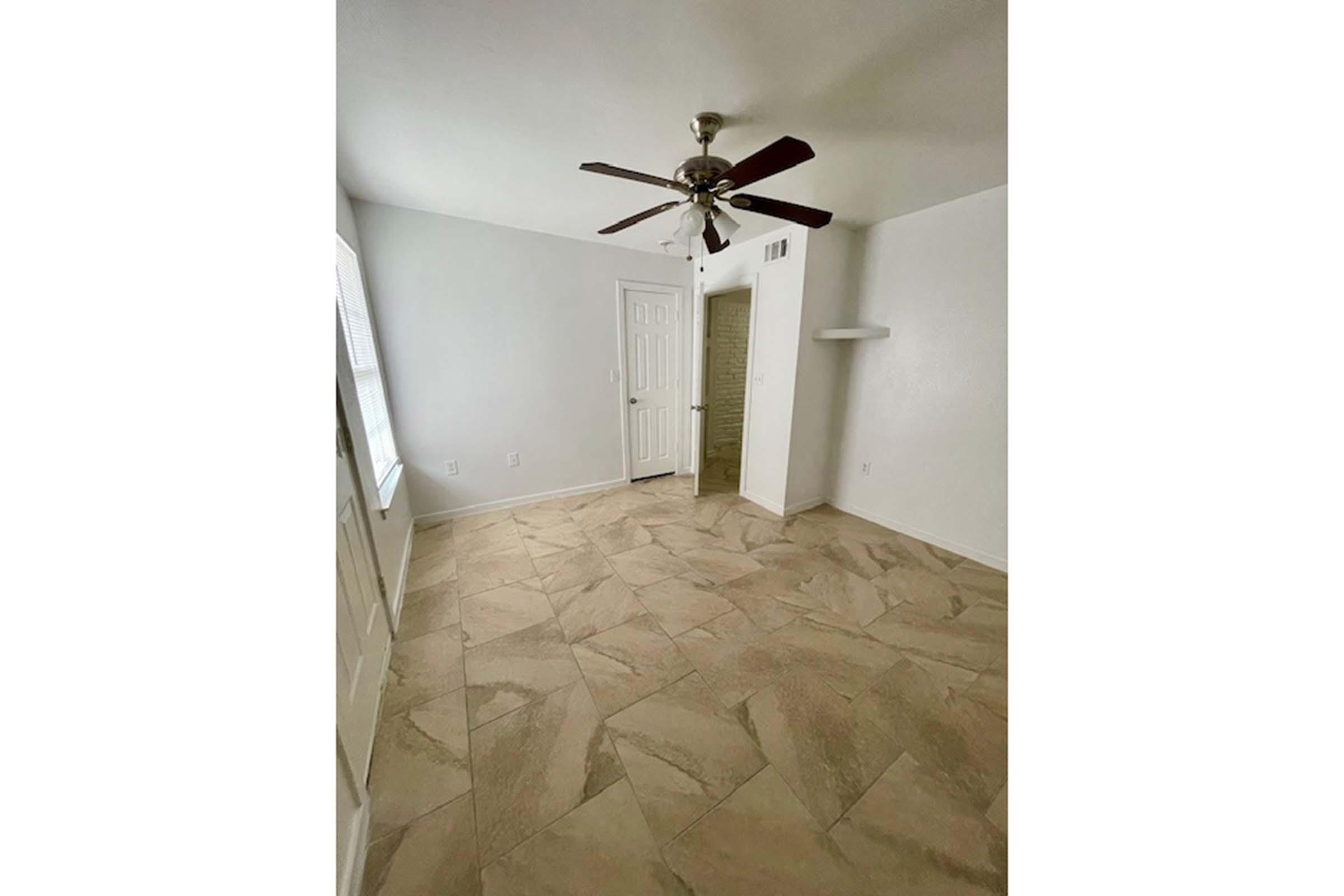 A spacious, empty room with light-colored walls and a tiled floor. It features a ceiling fan, a window with natural light, and a door leading to another area. A small shelf is mounted on one wall, and there's a closet space visible. The overall ambiance is bright and open.