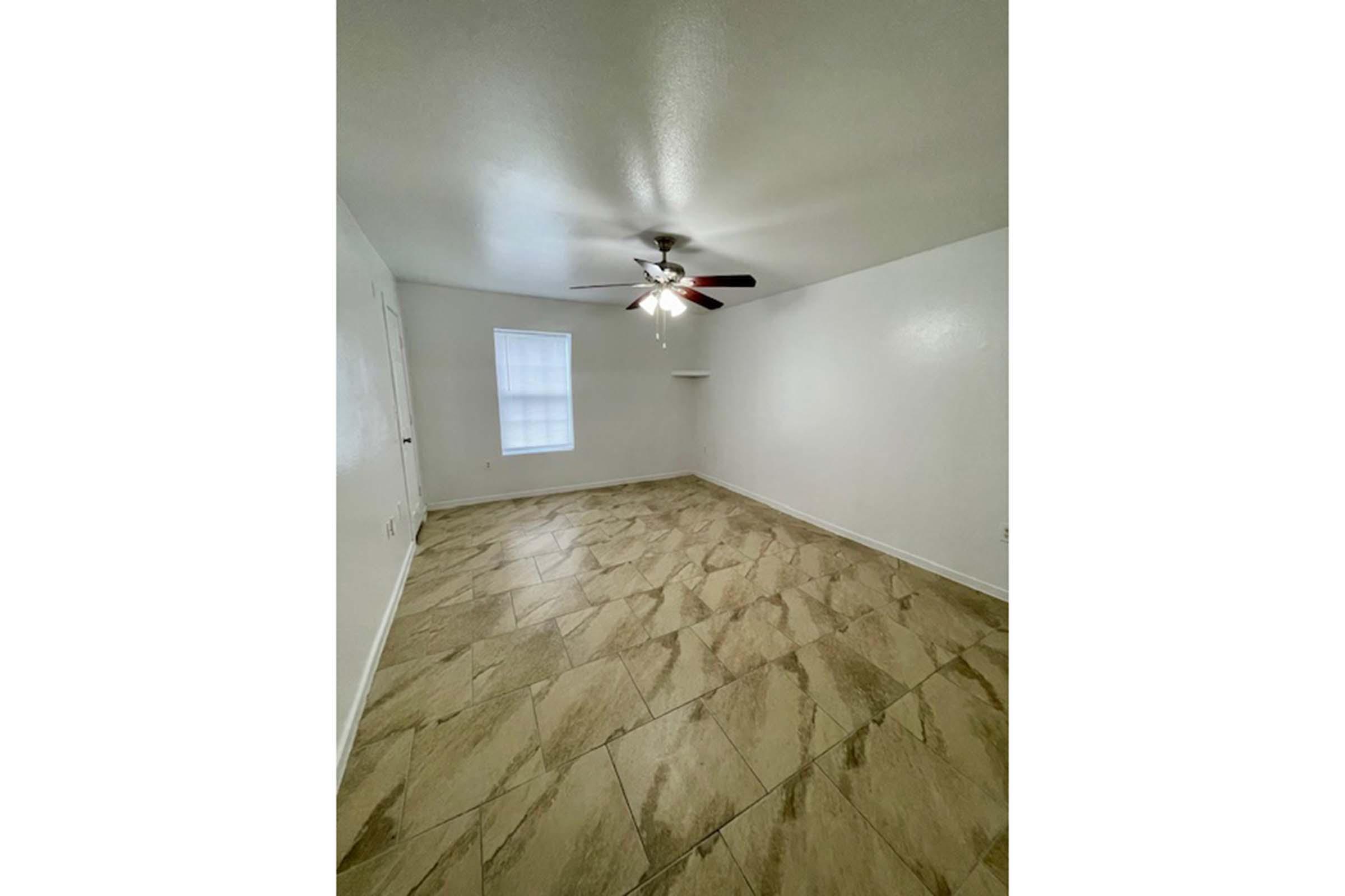 A spacious, empty room with beige tiled flooring, a ceiling fan with light, and a single window covered by sheer white curtains. The walls are painted white, and there is a small shelf on one side of the room. Natural light fills the space, creating a bright and airy atmosphere.