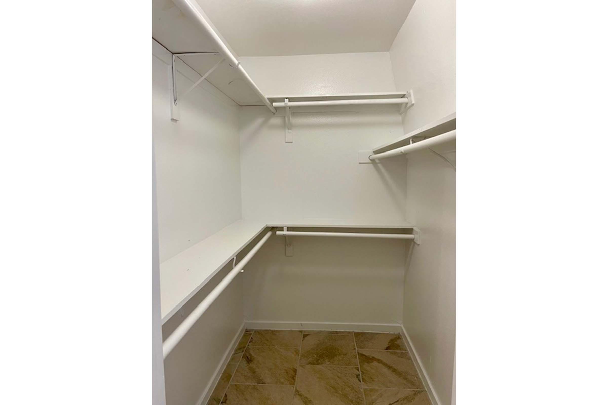 A clean, empty closet with white walls and tan tiled flooring. The closet features multiple white hanging rods and a shelf along one side, providing plenty of space for storage. The overall appearance is neat and minimalistic, making it suitable for organizing clothes or other items.