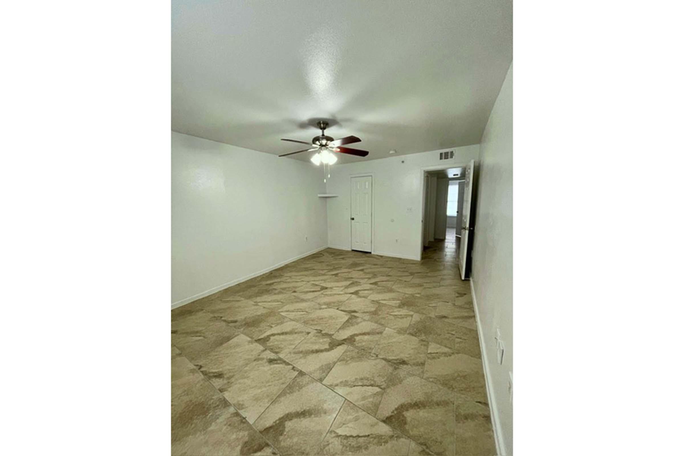 A spacious, empty room featuring light-colored walls and tile flooring. A ceiling fan is mounted in the center, and there are doors leading to other areas. The room is well-lit, with natural light coming from the open doorways.
